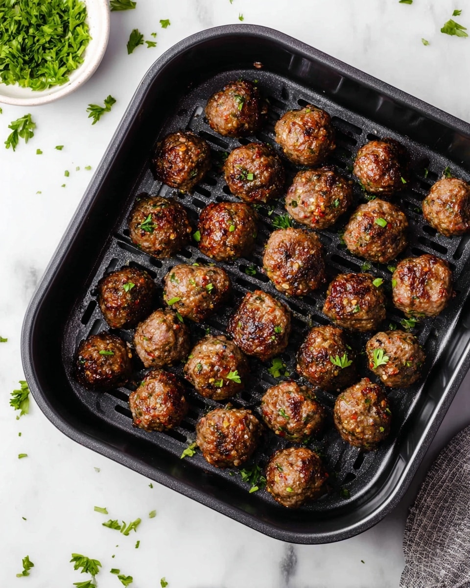 The image shows a black air fryer tray filled with 27 small, round meatballs that are browned and slightly shiny from cooking. The meatballs have a textured surface with bits of herbs and spices visible, and some green chopped parsley scattered over them and on the tray. The tray sits on a white marbled surface with some extra parsley pieces around it, and a small white bowl of green chopped herbs is partially visible in the top left corner. The overall look is clean and appetizing with a focus on the evenly cooked meatballs photo taken with an iphone --ar 4:5 --v 7