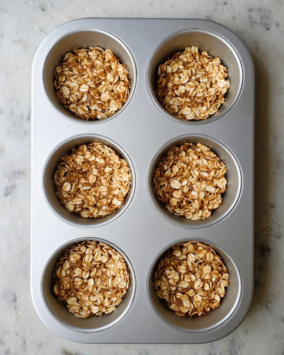 A silver muffin tray holds six cups, each filled with an oat mixture shaped like small bowls. The mixture looks sticky and textured, with visible oat flakes packed firmly to form hollow, round nests inside the shiny cups. The trays and oat nests rest on a white marbled surface, giving a clean and bright background. photo taken with an iphone --ar 4:5 --v 7