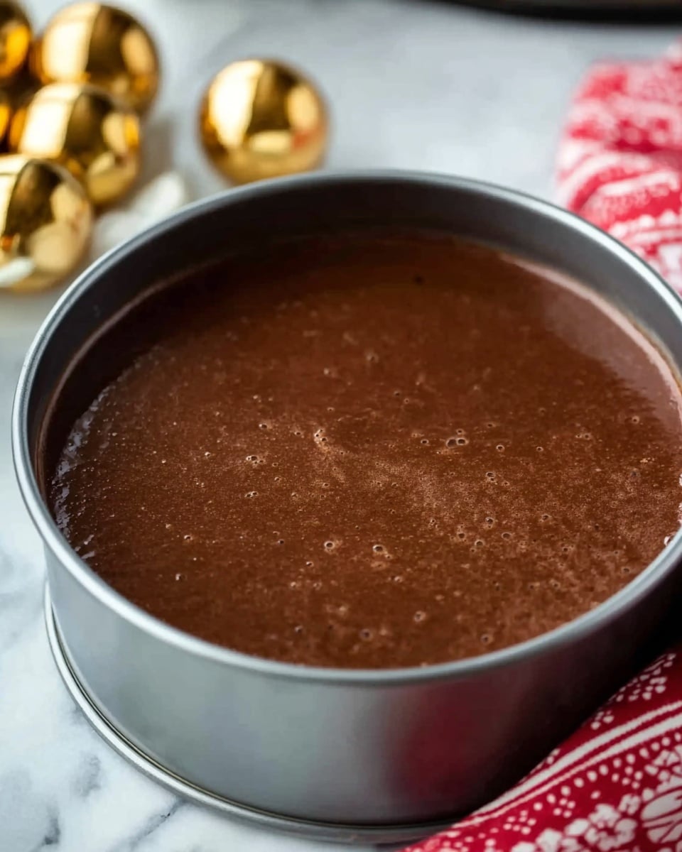 A close-up image of a round chocolate batter layer inside a metal cake ring, filled up to the top. The surface of the batter is smooth and shiny with a few small air bubbles, showing a rich dark brown color. The background is a white marbled texture, with golden decorative balls seen blurred on the left side and a red and white patterned cloth on the right side. Photo taken with an iphone --ar 4:5 --v 7