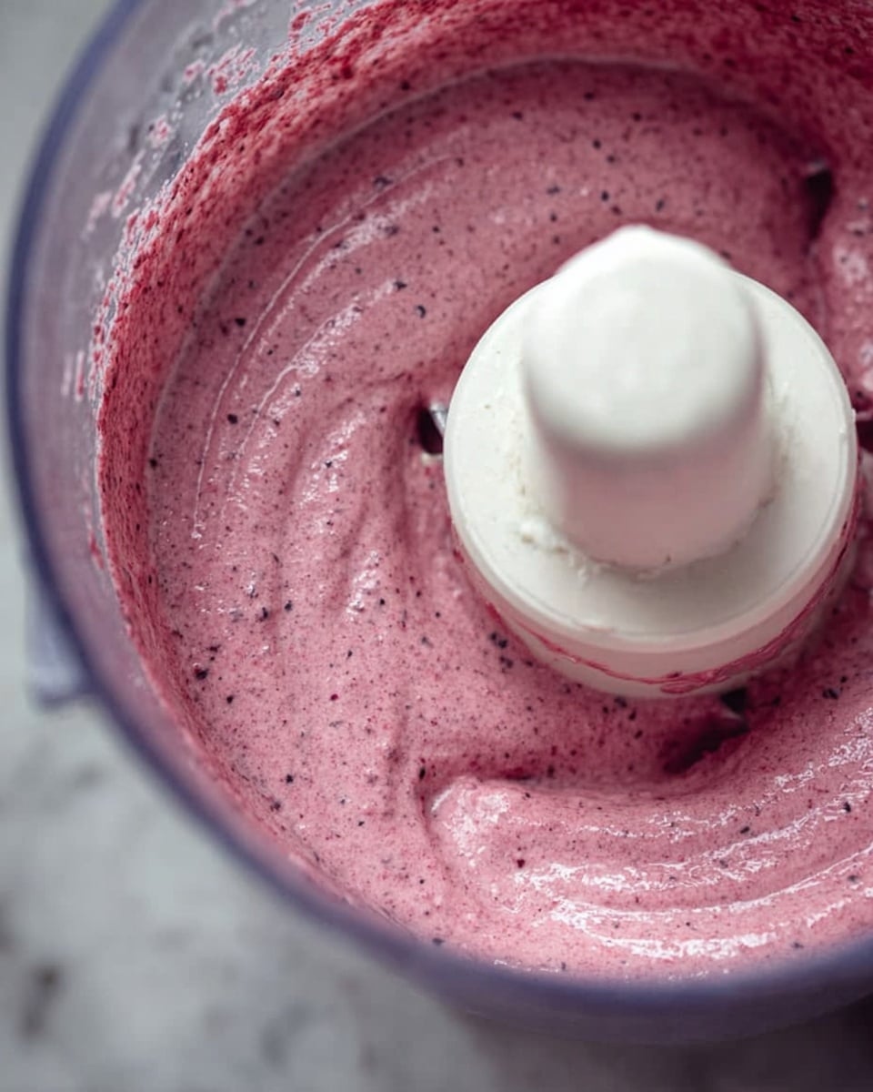 The image shows a close-up of a food processor bowl containing a thick, smooth pinkish-purple mixture with tiny dark specks, evenly spread around the white central blade attachment. The texture looks creamy and well-blended, with the mixture coating the base and sides of the transparent bowl. The background features a white marbled surface. photo taken with an iphone --ar 4:5 --v 7
