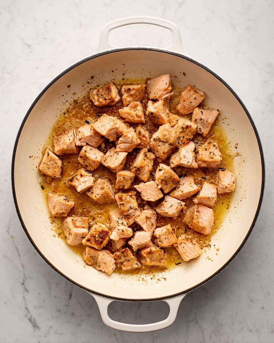 The image shows a top view of a white pan with two handles, placed on a white marbled surface. Inside the pan, there are many small, evenly chopped light pink and golden brown pieces of cooked salmon spread out in a single layer. The salmon pieces have a slightly flaky texture with some browned edges, and there is a thin layer of light golden brown cooking juices around them, with some darker spots where the fish has browned. The overall look is warm and inviting, showing the salmon ready to be served or used in a recipe. photo taken with an iphone --ar 4:5 --v 7