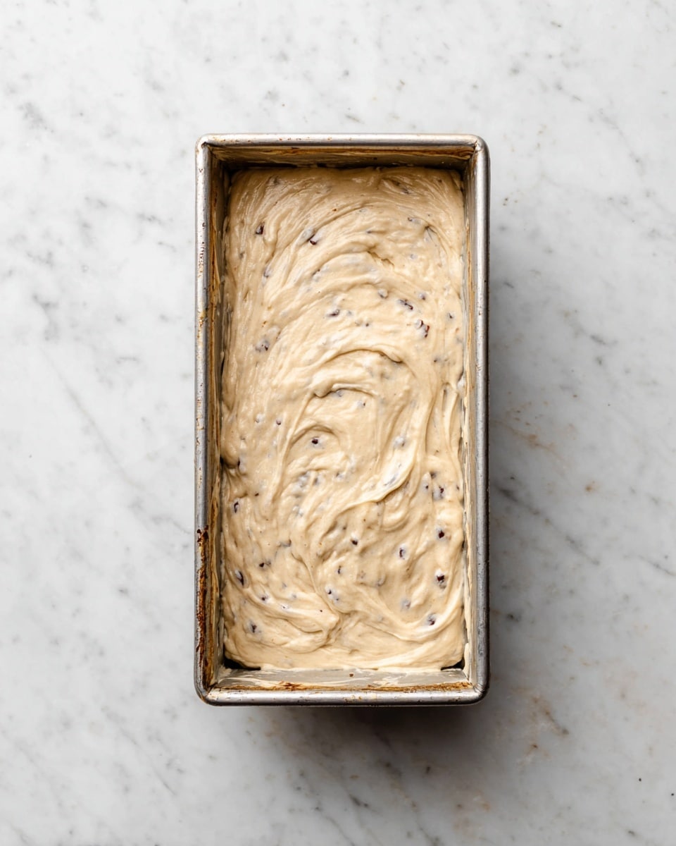A single rectangular metal baking pan filled with thick, creamy batter that is light beige with small dark specks mixed throughout. The batter is spread in uneven swirls and covers the pan evenly. The pan sits on a white marbled surface, showing some wear and slight browning around the edges inside the pan. photo taken with an iphone --ar 4:5 --v 7