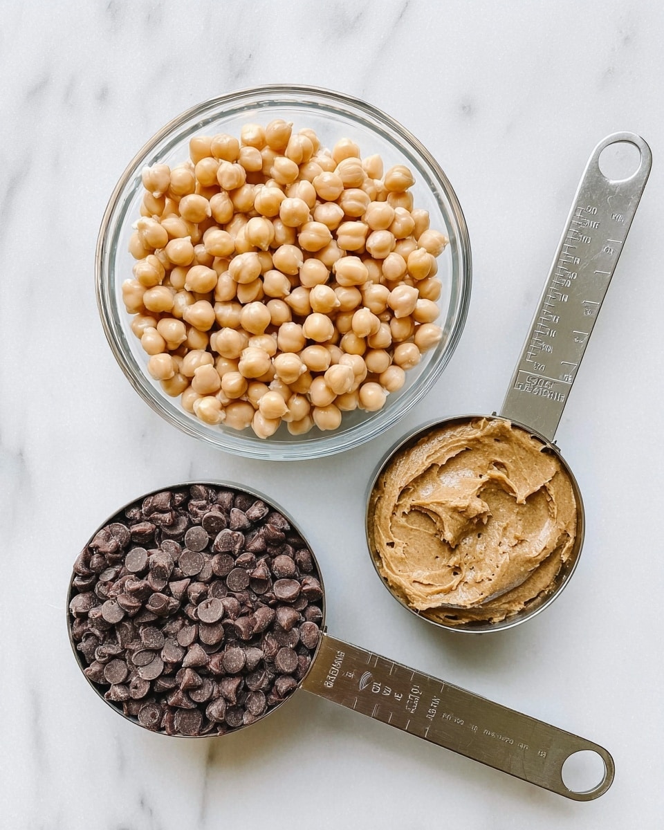 The image shows three containers with different food items arranged on a white marbled surface. At the top center, there is a clear glass bowl filled with light beige chickpeas, each round and smooth. Below it, slightly to the left, is a silver measuring cup filled to the brim with small, dark brown chocolate chips, which have a shiny and slightly curved texture. To the right of the chocolate chips, there is another silver measuring cup holding a light brown, creamy nut butter, smooth in texture with some small darker specks visible. The handles of the measuring cups have measurement markings visible. photo taken with an iphone --ar 4:5 --v 7