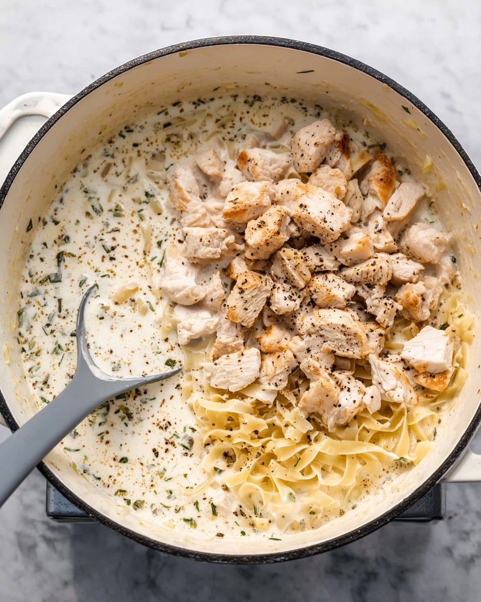 A large white pot is filled with creamy white sauce that has black pepper and green herbs sprinkled throughout. At the bottom of the pot, there are thin, flat noodles partly submerged in the sauce. On top of the sauce and noodles, there is a pile of chopped light brown cooked chicken pieces. A gray spoon with a short handle is partly dipped into the sauce on the left side of the pot. The pot is sitting on a white marbled surface. photo taken with an iphone --ar 4:5 --v 7