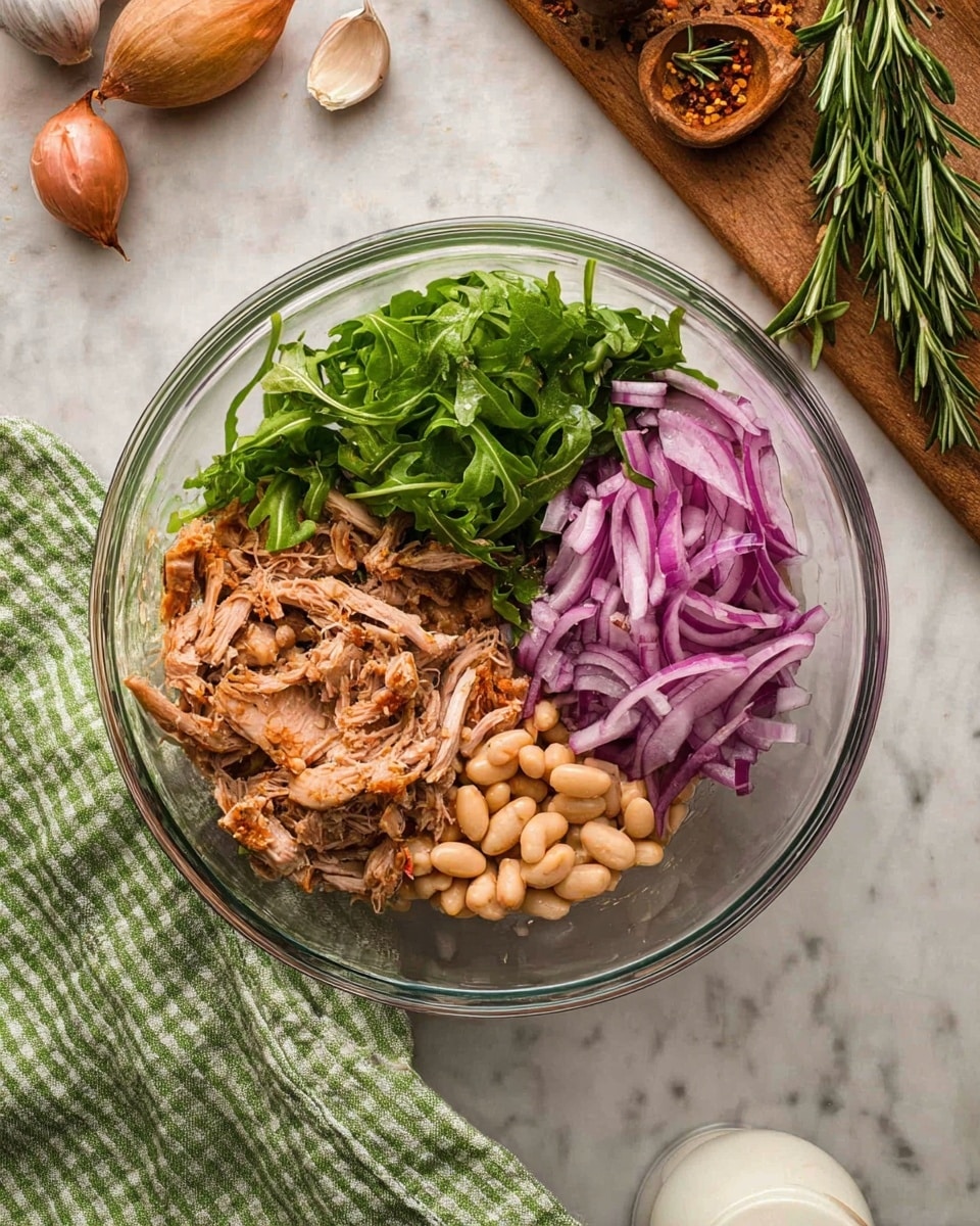 A clear glass bowl is filled with four distinct layers of ingredients arranged side by side inside: shredded light brown roasted meat with spices at the bottom right, thin slices of purple-red onion on the top right, light beige beans in the center, and fresh green arugula leaves on the left side; the bowl sits on a white marbled surface with a green and white striped cloth draped nearby, and in the background are whole garlic cloves, a shallot, and some rosemary on a wooden board, with a small white container partially visible at the bottom right, photo taken with an iphone --ar 4:5 --v 7