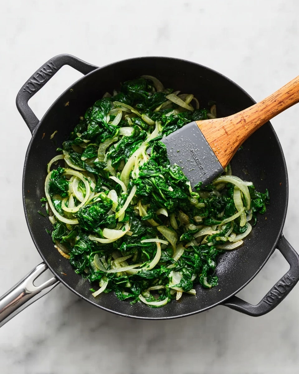 In a black pan with two handles, bright green cooked spinach and pale translucent onion slices are mixed together. The spinach forms clumps along the bottom, with round onion rings scattered evenly throughout. A wooden spatula with a gray rubber head rests inside the pan, slightly lifting some of the spinach and onions. The pan sits on a white marbled surface, clean and bright. photo taken with an iphone --ar 4:5 --v 7