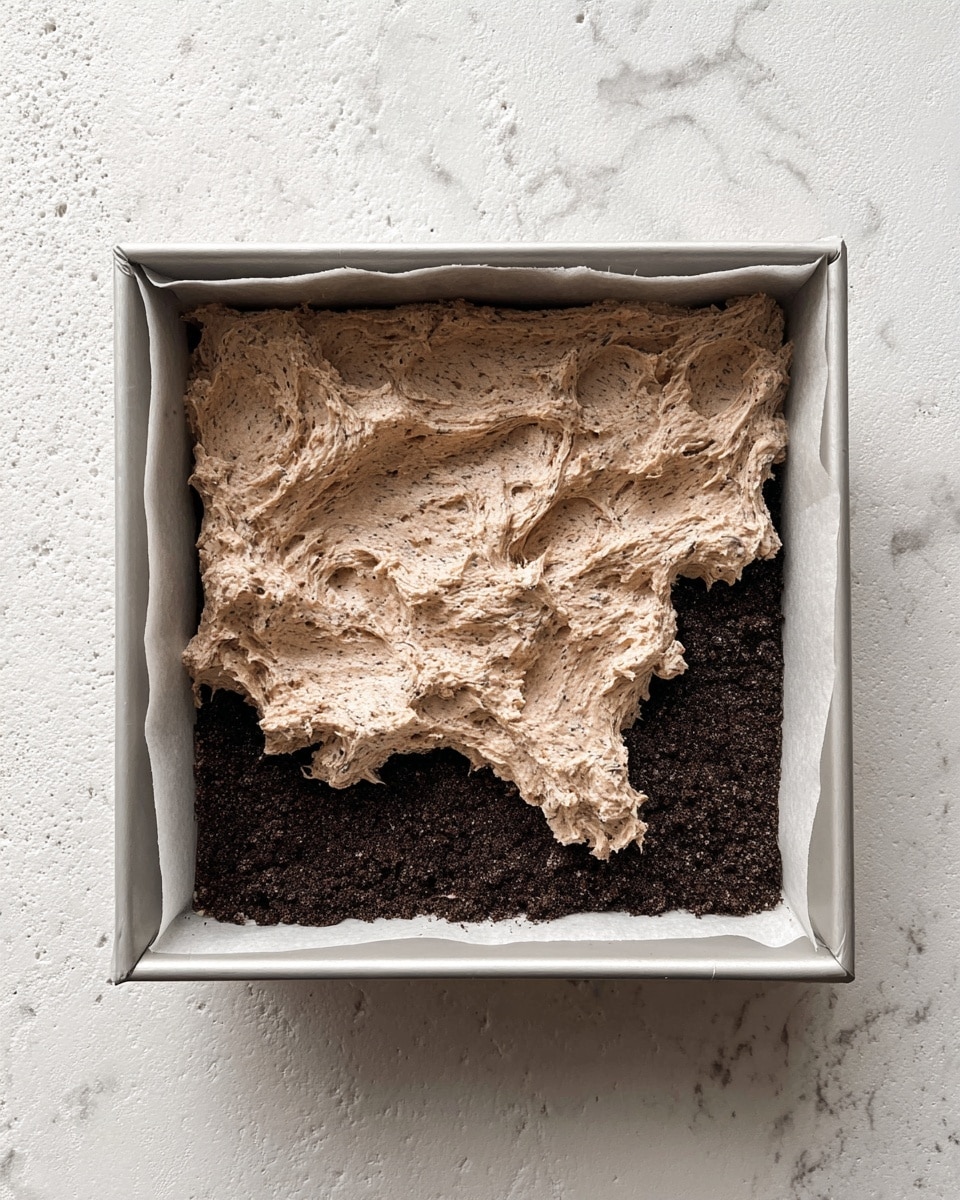 A square metal baking pan lined with white parchment paper holds two visible layers: the bottom layer is dark brown, finely crushed cookie crumbs pressed flat to form a solid base, while the top layer is a thick, uneven spread of light brown creamy mixture with small dark specks, giving it a textured, whipped appearance that covers most of the base but not evenly. The pan is placed on a white marbled textured surface. photo taken with an iphone --ar 4:5 --v 7