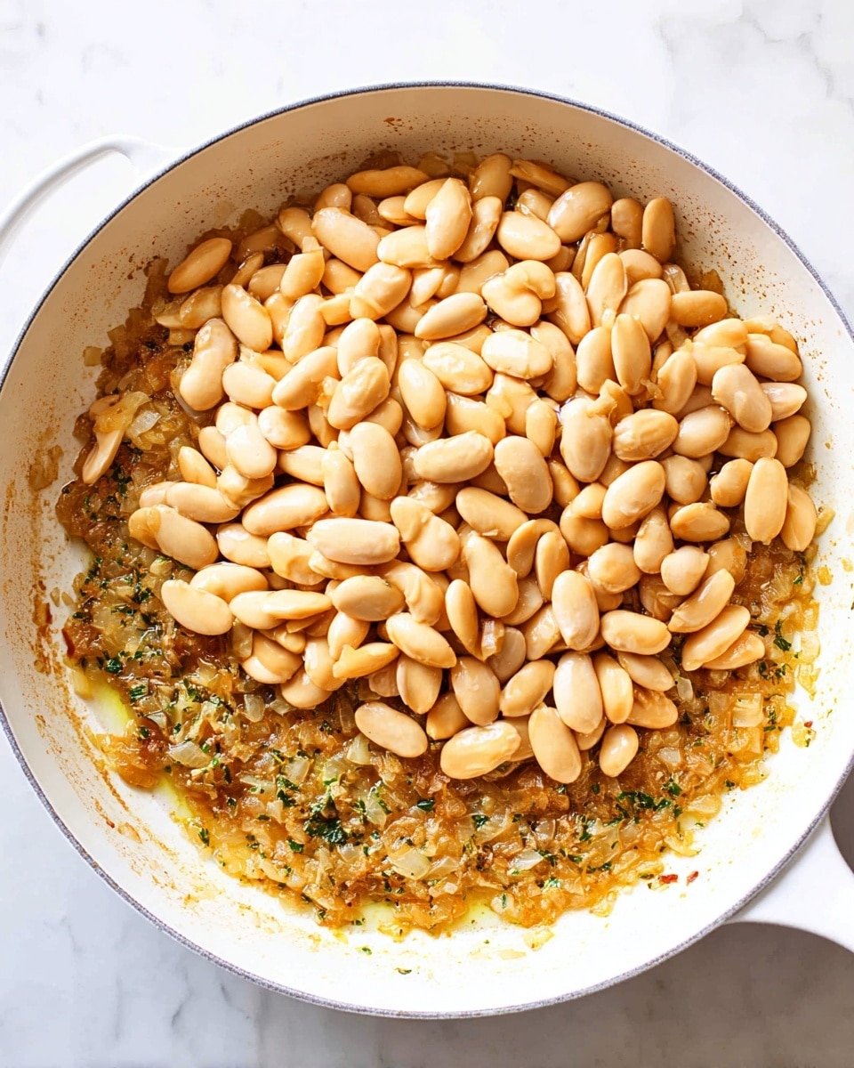 A white pan filled with two layers: the bottom layer is finely chopped cooked onions mixed with herbs and oil, showing a golden brown texture and some visible herbs, while the top layer consists of a thick pile of whole light tan beans resting on the onion mixture. The pan sits on a white marbled surface. photo taken with an iphone --ar 4:5 --v 7