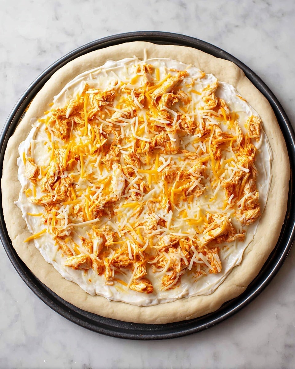 A pizza with three layers is shown on a black round tray. The bottom layer is a thick, pale dough crust with a slightly puffy edge. The second layer is a smooth, creamy white sauce spread evenly on the dough. The top layer has a mix of shredded yellow and white cheese scattered across the sauce, with pieces of orange-coated shredded chicken spread over the cheese. The pizza is placed on a white marbled surface. photo taken with an iphone --ar 4:5 --v 7