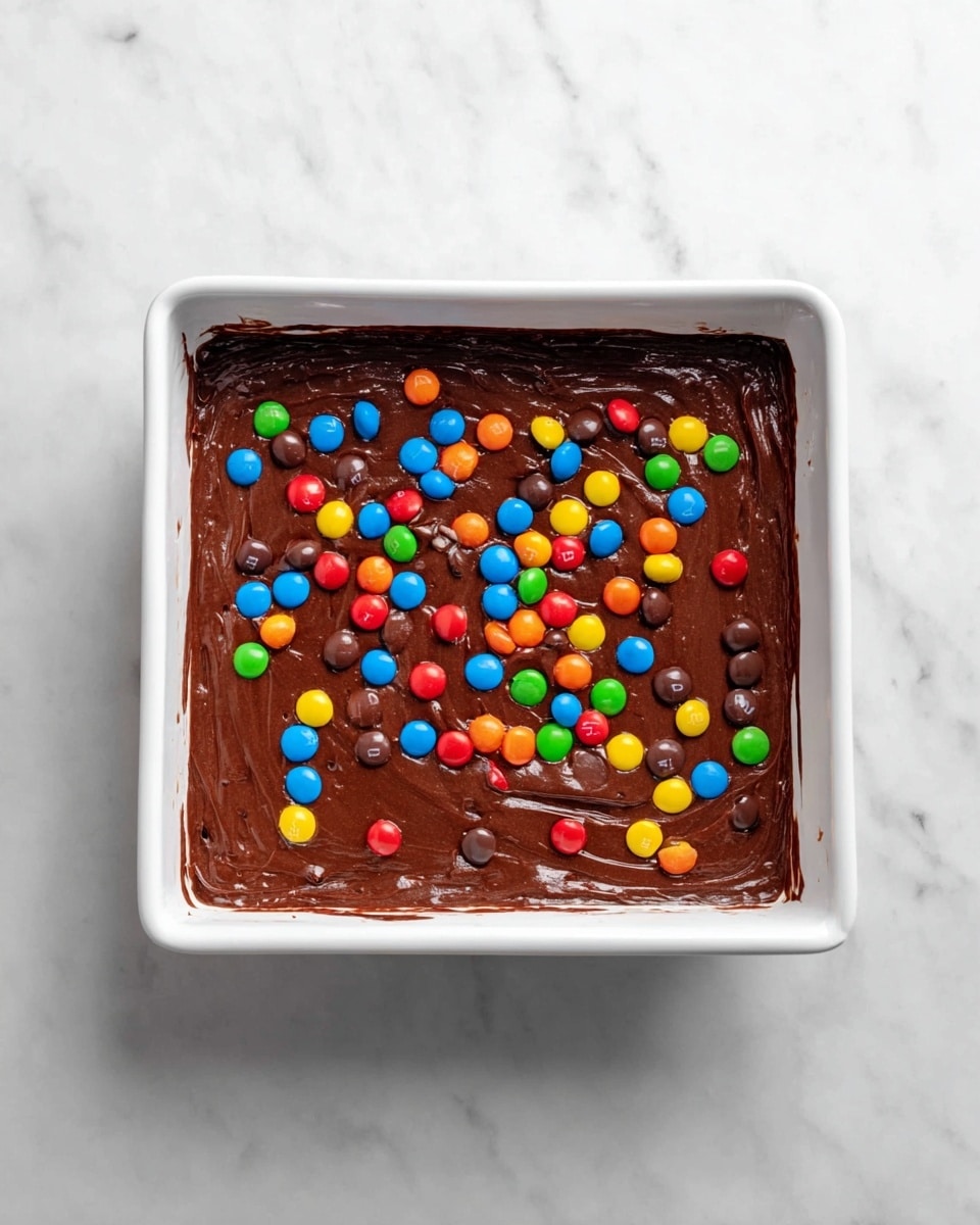 The image shows a white square baking pan filled with a thick layer of smooth, dark brown chocolate batter spread evenly across the bottom. On top of the chocolate batter, there is a scattered layer of colorful candy-coated chocolates in bright red, blue, green, yellow, orange, and brown, adding a lively contrast to the dark batter. The pan rests on a clean white marbled surface, giving the scene a fresh and bright look. photo taken with an iphone --ar 4:5 --v 7