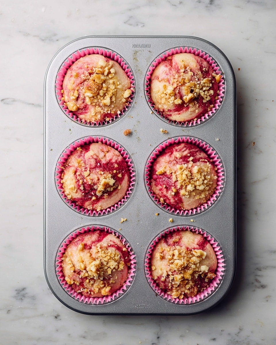 A silver muffin tray holds six unbaked muffins, each in a pink paper liner. The muffins are a mix of light pink and red swirls, showing a marbled texture with bits of crushed nuts sprinkled on top. The tray sits on a white marbled surface, creating a clean and bright background. Photo taken with an iphone --ar 4:5 --v 7