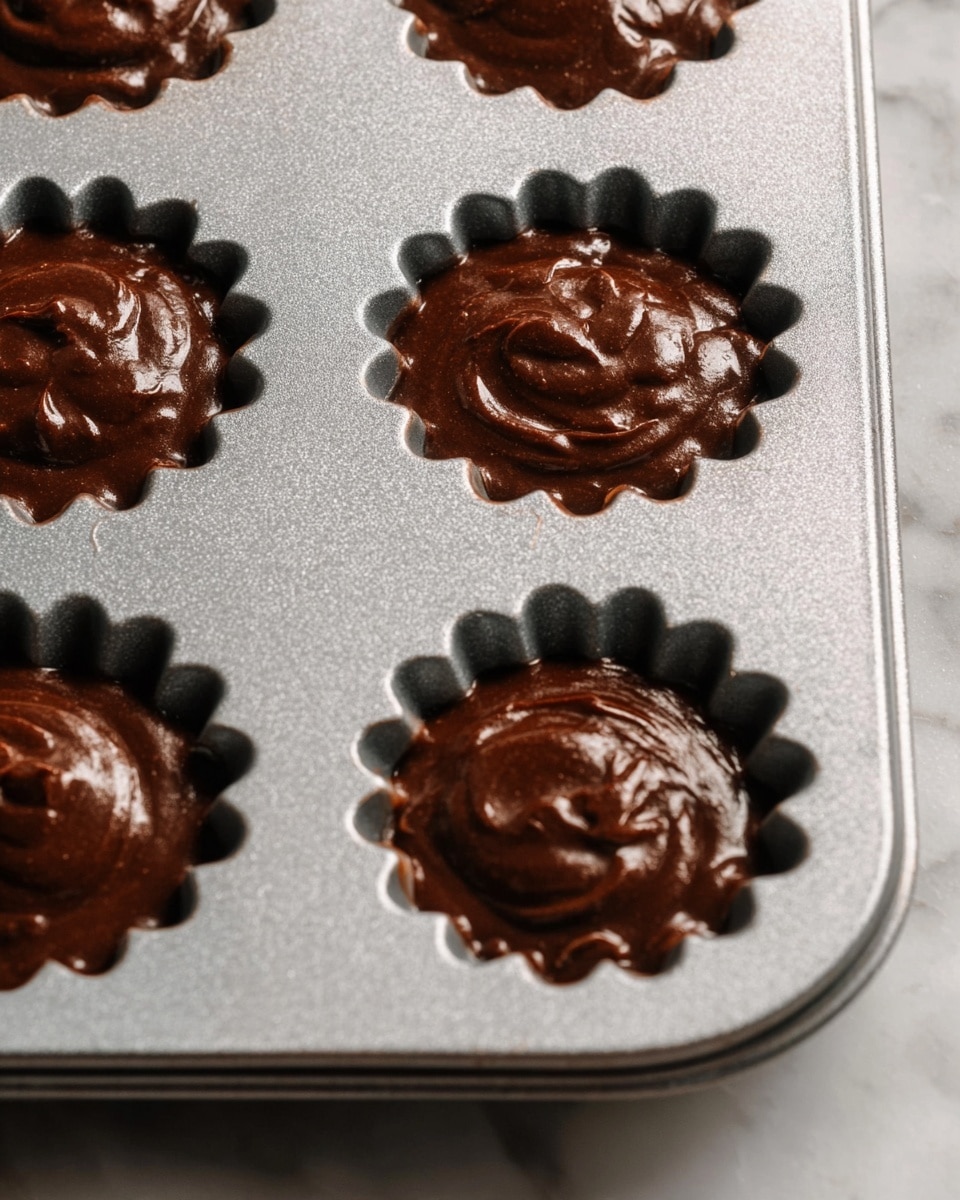 A close-up view of a silver metal baking tray filled with chocolate batter in six visible flower-shaped molds. Each mold is filled with a rich, thick, dark brown chocolate batter with a smooth and slightly swirled texture. The tray sits on a white marbled surface. photo taken with an iphone --ar 4:5 --v 7