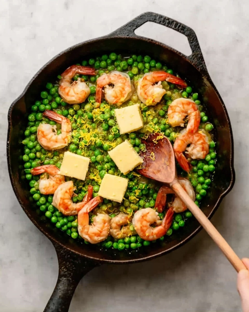 A black cast iron pan filled with a layer of bright green peas covering the bottom. On top of the peas, there are cooked shrimp arranged evenly around the pan, showing their orange and pink color with light grill marks. Near the shrimp, there are two square pieces of yellow butter melting, with some yellow lemon zest sprinkled across the peas and shrimp. A woman's hand holds a wooden spoon resting inside the pan. The background surface is white marbled. photo taken with an iphone --ar 4:5 --v 7