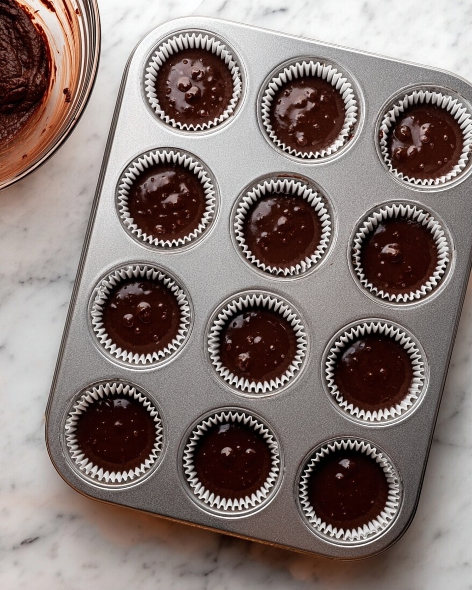 A metal cupcake tray with twelve slots is shown filled with dark brown chocolate batter inside white cupcake liners. The batter has a shiny, smooth texture with small visible bubbles on the surface, filling each liner about three-quarters full. The tray is placed on a white marbled surface, and on the left edge of the image, there is a glass bowl with some chocolate batter remnants around the edge. Photo taken with an iphone --ar 4:5 --v 7