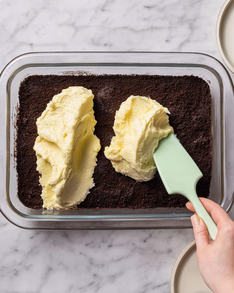 A glass rectangular dish with a dark brown crumb layer spread evenly at the bottom fills most of the frame. On top, there are two large dollops of thick, creamy, light yellow mixture placed near the center and right side of the dish. A woman's hand is holding a pale green spatula spreading the creamy mixture over the dark crumb base. The dish sits on a white marbled surface with part of a white plate visible on the right side. Photo taken with an iphone --ar 4:5 --v 7