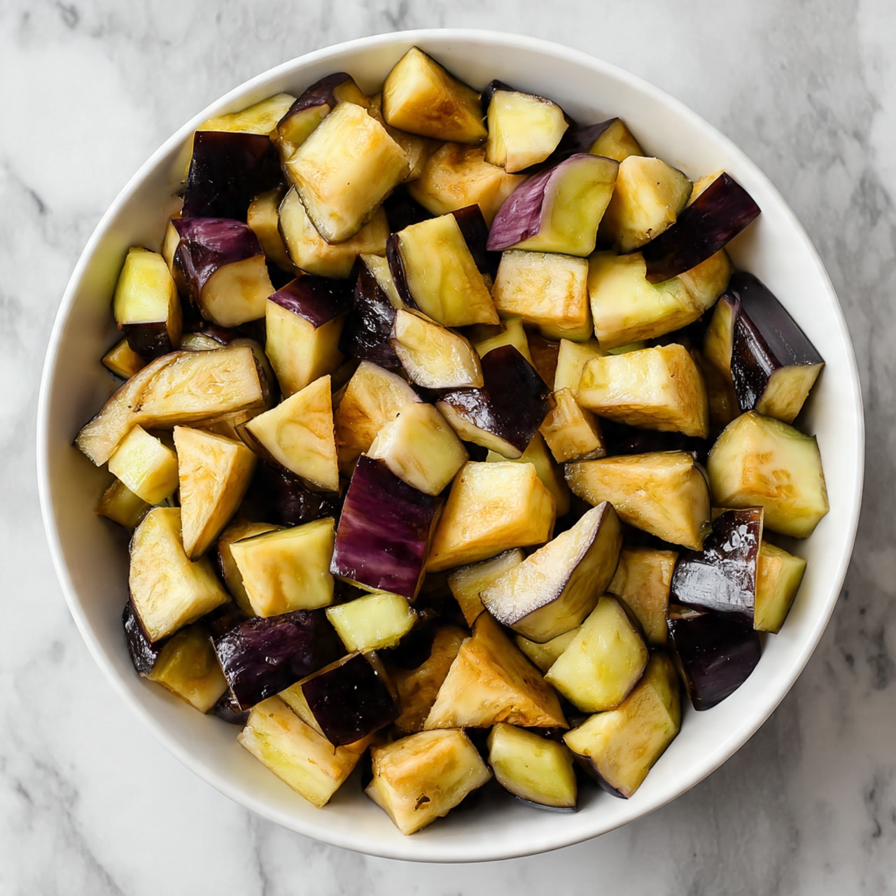 A round white bowl filled with many pieces of chopped eggplant. The eggplant pieces have dark purple skin edges and light yellowish flesh. Some pieces are triangular or rectangular in shape. The eggplant looks seasoned with a light coating that gives the flesh a soft texture. The bowl is placed on a white marbled surface. photo taken with an iphone --ar 4:5 --v 7