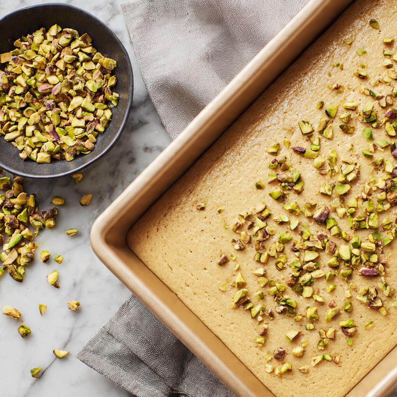 A close-up view of a rectangular beige baking pan filled with a light tan, smooth mixture that looks soft and slightly grainy. On top of the mixture, small pieces of chopped green and brown pistachios are scattered unevenly. To the left of the pan, there is a small black bowl filled with more chopped pistachios, with some pieces spilling out onto the white marbled surface underneath. The background includes a soft gray cloth with faint folds visible. photo taken with an iphone --ar 4:5 --v 7