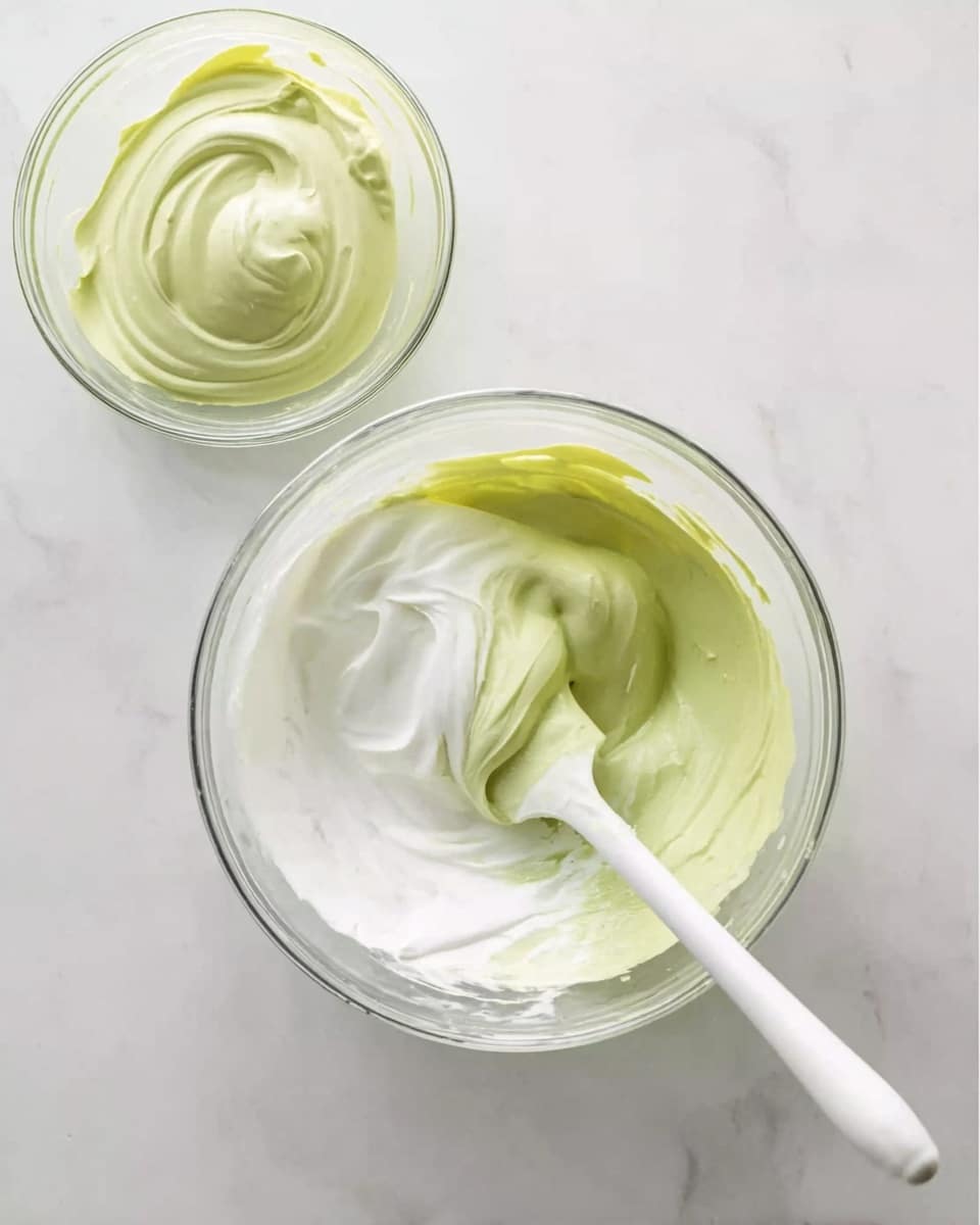 A clear glass bowl sits on a white marbled surface, containing two swirled layers of creamy mixture—one layer light green and smooth, and the other white and fluffy—gently combined with a white spatula whose handle leans inside the bowl. Above this bowl, in the top left corner of the image, is a smaller clear bowl filled with more of the light green creamy layer, showing its thick, smooth texture. The overall look is soft and fresh with a calm, clean background. Photo taken with an iphone --ar 4:5 --v 7