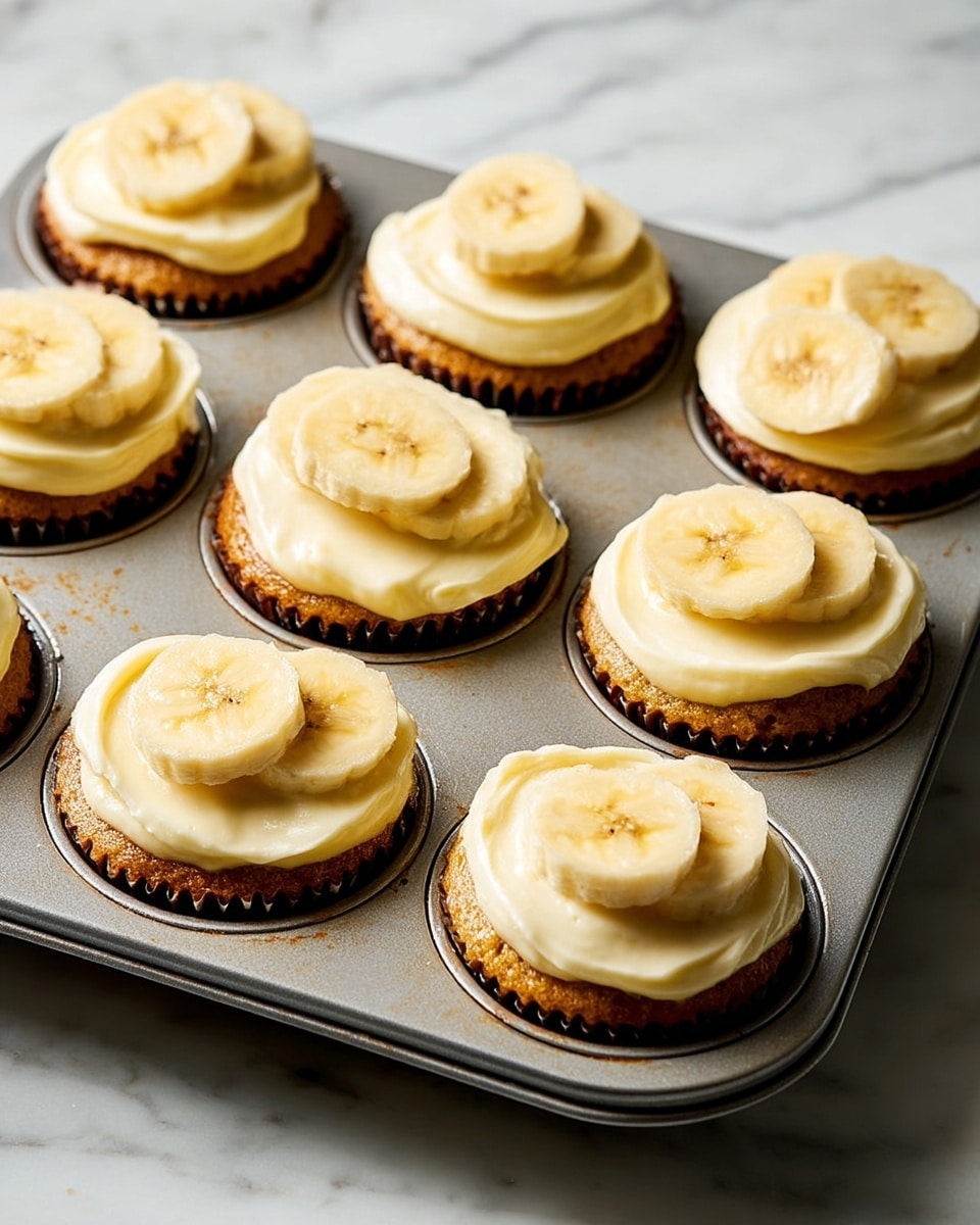A metal muffin tray holds nine cupcakes, each with three layers: the bottom is a golden brown cupcake base in dark paper liners, the middle is a thick layer of smooth, pale yellow frosting, and the top is decorated with three to four banana slices that appear soft and fresh. The tray sits on a white marbled surface, and the light highlights the creamy texture of the frosting and the subtle shine on the banana slices. photo taken with an iphone --ar 4:5 --v 7
