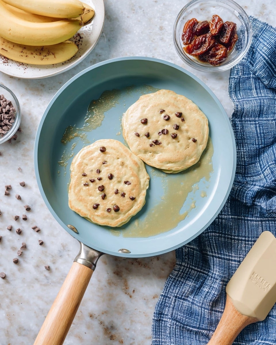 Inside a light blue nonstick frying pan, two thick, pale yellow pancake-like batter discs with small dark chocolate chips are cooking, sizzling with spots of melted butter around them. The pan rests on a white marbled surface, with a blue checkered cloth and a beige spatula with a wooden handle beside it. Nearby, a small white bowl contains dark chocolate chips scattered slightly outside, a clear glass dish holds chopped dates, and a white plate shows sliced banana pieces. Photo taken with an iphone --ar 4:5 --v 7