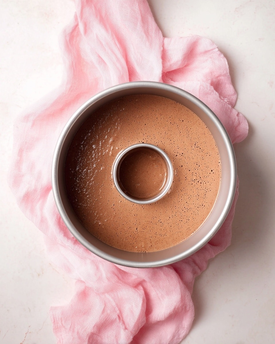 A round silver bundt cake pan filled with smooth, light brown chocolate batter that has small bubbles on the surface. The pan is placed on top of a soft pink cloth that lies on a white marbled surface. The image is shot from above, showing the even layer of batter inside the pan. photo taken with an iphone --ar 4:5 --v 7