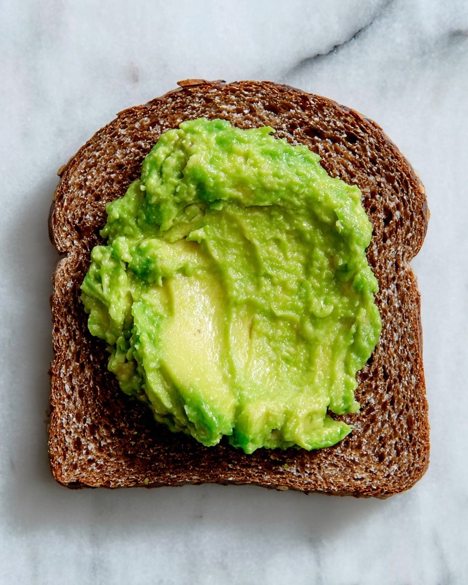 A single slice of dark brown bread with a coarse texture lies flat on a white marbled surface. On top, near the center, there is a thick dollop of bright green avocado spread with a smooth but slightly chunky texture. The light and shadow highlight the contrast between the rough bread and the creamy avocado photo taken with an iphone --ar 4:5 --v 7