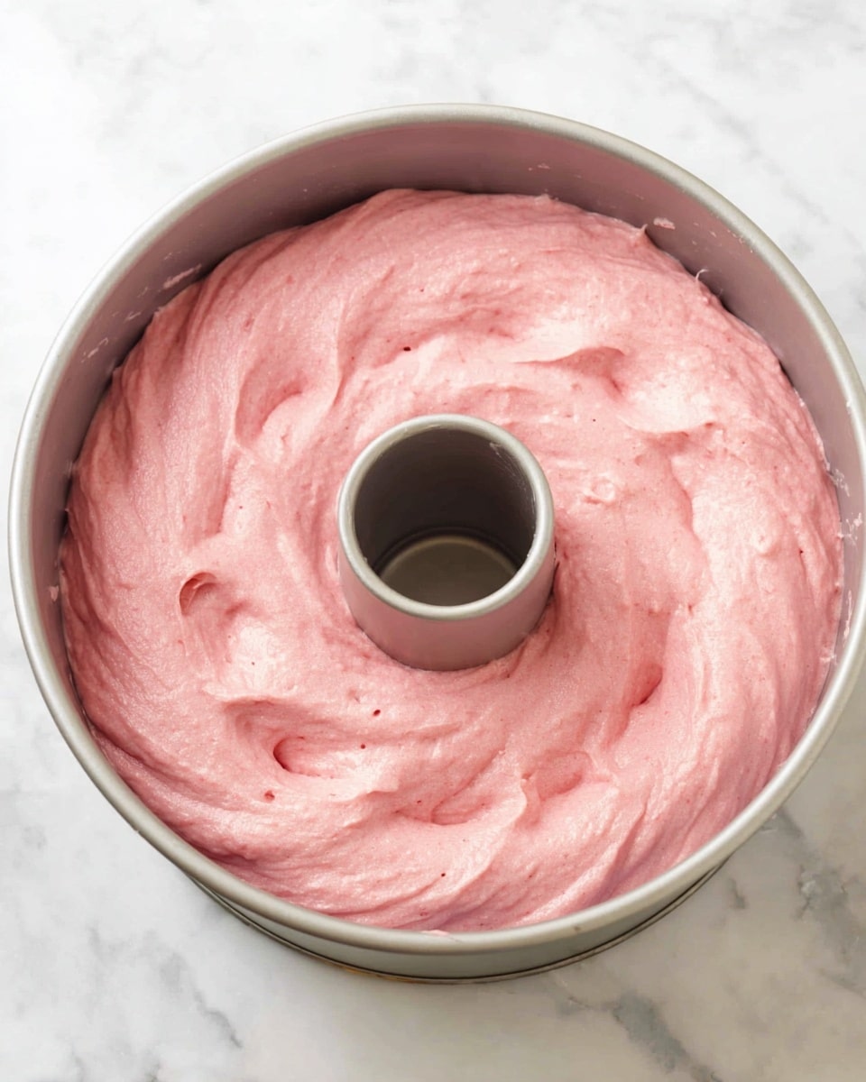 The image shows a light gray metal bundt pan filled with smooth, thick pink cake batter spread evenly to the edges and around the central tube, covering the entire pan surface in one layer. The batter has a creamy texture with soft swirls and small air pockets, giving it a light, fluffy look. The pan is placed on a white marbled surface, creating a clean and bright background. Photo taken with an iphone --ar 4:5 --v 7