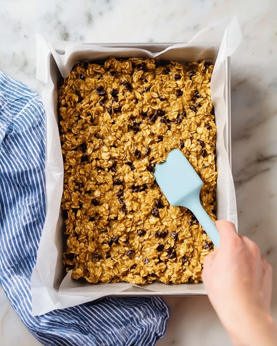 A clear glass bowl contains three layers: the bottom layer is a smooth yellowish batter, the top left half is covered with pale beige rolled oats, and the top right half has a large pile of dark brown chocolate chips. A light blue spatula rests inside the bowl, its blade touching the batter, oats, and chocolate chips. The bowl is placed on a white marbled surface, surrounded by three empty white bowls and a small clear glass of water, with a blue and white striped cloth on the left side. photo taken with an iphone --ar 4:5 --v 7