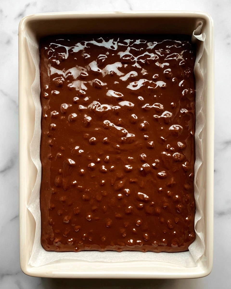 The image shows a single layer of thick, glossy chocolate batter filled with small, raised nut or chip pieces spread evenly inside a white rectangular baking dish lined with parchment paper. The surface of the batter is smooth with shiny highlights reflecting light, and the texture looks rich and dense with visible rounded bits giving it a bumpy appearance. The dish is placed on a white marbled surface. Photo taken with an iphone --ar 4:5 --v 7