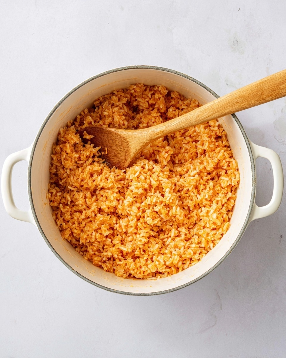 A white pot filled with orange-colored cooked rice with visible grain texture. Inside the pot, a wooden spoon is placed with its handle extending outward. The pot has two short handles on each side and is set on a white marbled surface. The rice looks soft and evenly cooked with a slightly sticky appearance. photo taken with an iphone --ar 4:5 --v 7