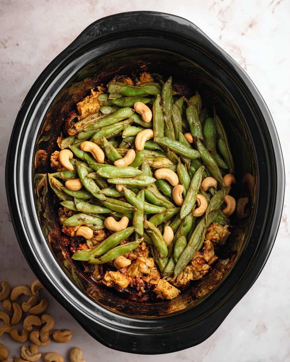 Inside a black slow cooker, there are three visible layers. The bottom layer is a dark, slightly charred base. On top of it, there are golden-brown cooked pieces with a rough texture, mixed with whole cashew nuts that are light beige and smooth. The top layer is made of many green snap peas that have a slightly wrinkled texture and cover the dish evenly. The slow cooker sits on a white marbled surface with scattered cashew nuts to the side. photo taken with an iphone --ar 4:5 --v 7
