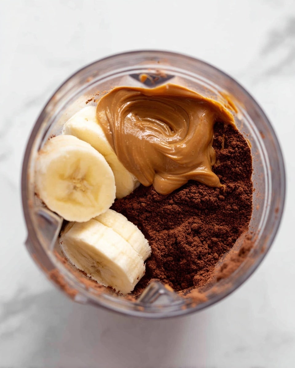 Inside a clear blender jar, the bottom layer shows a rich, dark brown cocoa powder with a soft, powdery texture. On top of this, three thick slices of pale yellow banana rest, their smooth and creamy appearance forming the second layer. Poured over the banana slices is a thick swirl of caramel-brown nut butter, glossy and slightly dripping onto the fruit. The jar is placed on a white marbled surface, and the image is captured from a top-down view. photo taken with an iphone --ar 4:5 --v 7