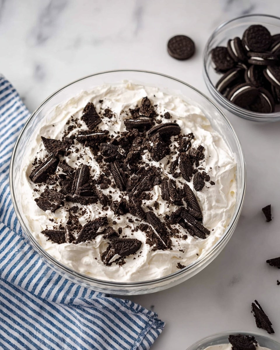 A clear glass bowl filled with a thick, white whipped cream layer topped with many broken dark chocolate sandwich cookie pieces scattered unevenly on the surface. In the top left, another glass bowl contains more broken cookies, showing dark brown and white layers. To the top right, a blue and white striped cloth rests on a smooth white marbled surface, which forms the background of the image. photo taken with an iphone --ar 4:5 --v 7