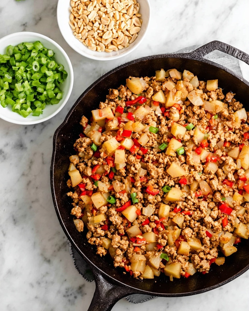 A black cast iron skillet filled with a cooked mixture of finely chopped pale beige ground meat, small chunks of light yellow potato, and diced red bell pepper pieces, all mixed closely together with a slightly glossy texture. Above the skillet, there are two small white bowls, one filled with bright green chopped spring onions and the other with crushed pale beige peanuts. The skillet and bowls are placed on a white marbled surface. photo taken with an iphone --ar 4:5 --v 7