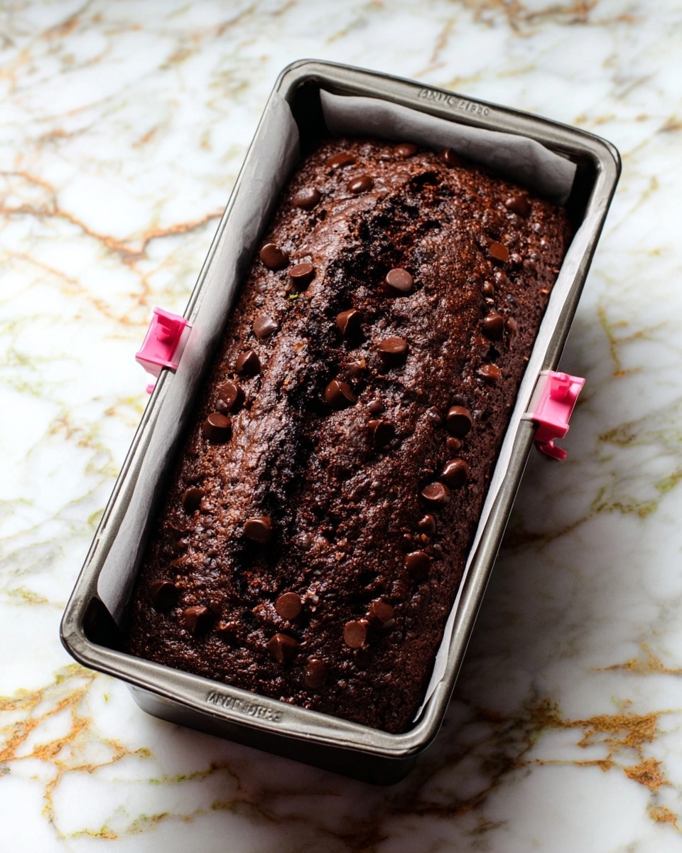A dark brown chocolate cake loaf with a cracked, rough top layer covered in small round chocolate chips scattered evenly. The cake is inside a metal baking pan lined with white parchment paper clipped on the sides with pink clips. The pan sits on a white marbled surface with veins of grey and soft gold running through it. Photo taken with an iphone --ar 4:5 --v 7