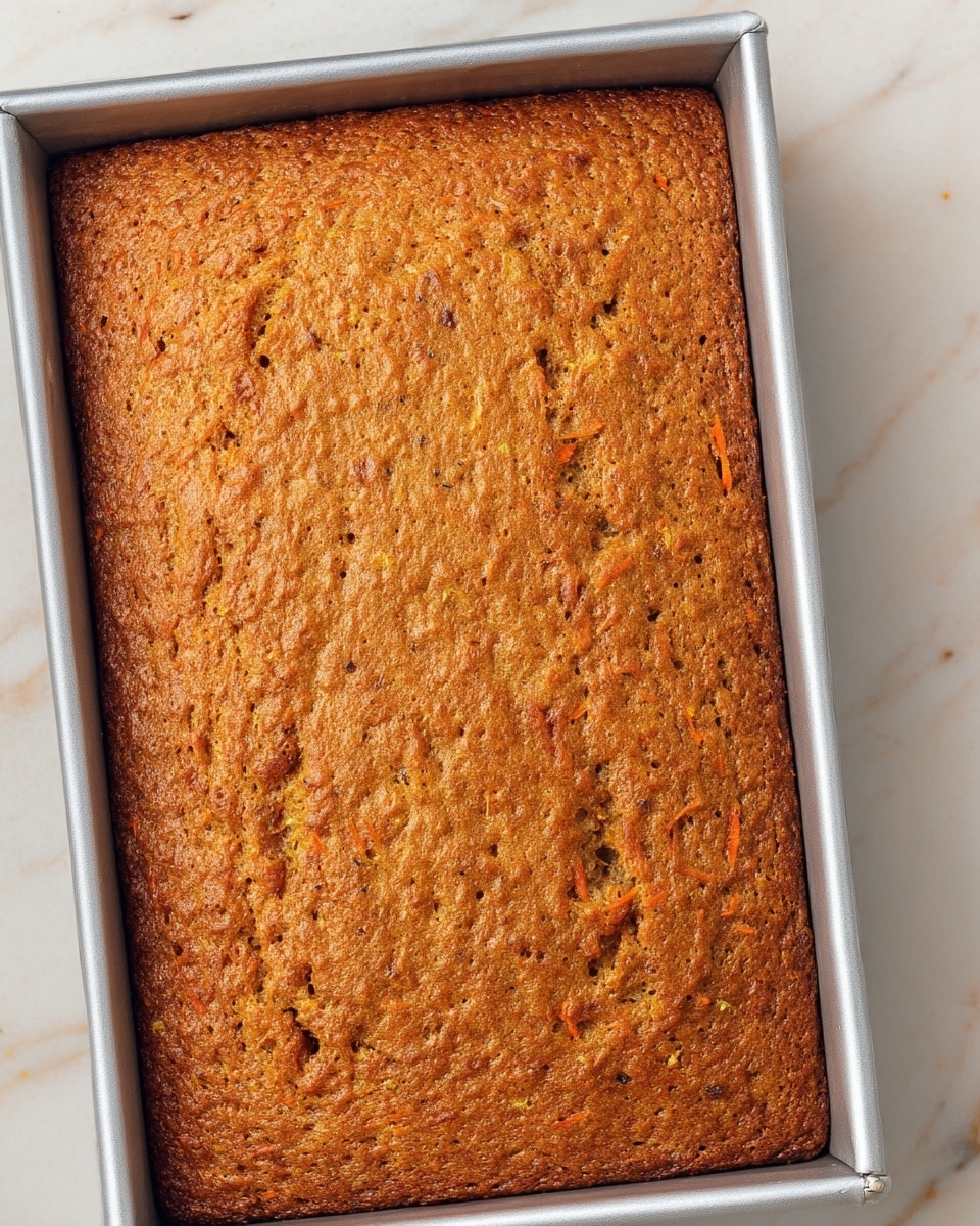A rectangular baked carrot cake with one thick layer fills a silver baking pan sitting on a white marbled surface. The cake's top is a warm brown color with visible small carrot shreds and tiny dark specks throughout, indicating spices or nuts. The texture looks firm but soft, with a slightly uneven surface showing natural baking bubbles and a golden-brown crust. Photo taken with an iphone --ar 4:5 --v 7
