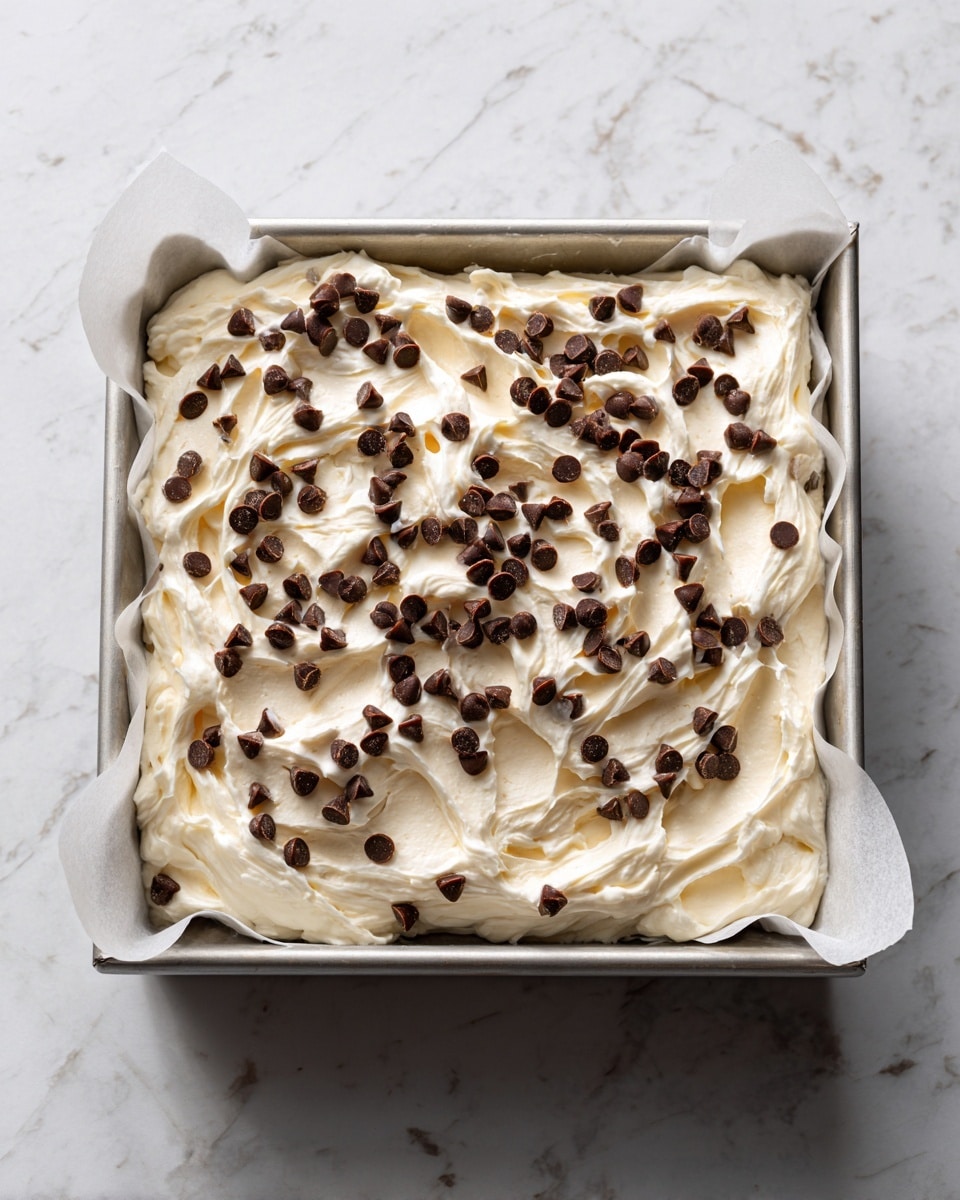 A square metal baking pan lined with white parchment paper holds one thick layer of white, creamy dough mixed with small dark chocolate chips and chopped green pistachios scattered evenly on top and through the dough. The surface beneath the pan has a white marbled texture. The number 4 in black is inside a white circle near the top left corner of the image photo taken with an iphone --ar 4:5 --v 7