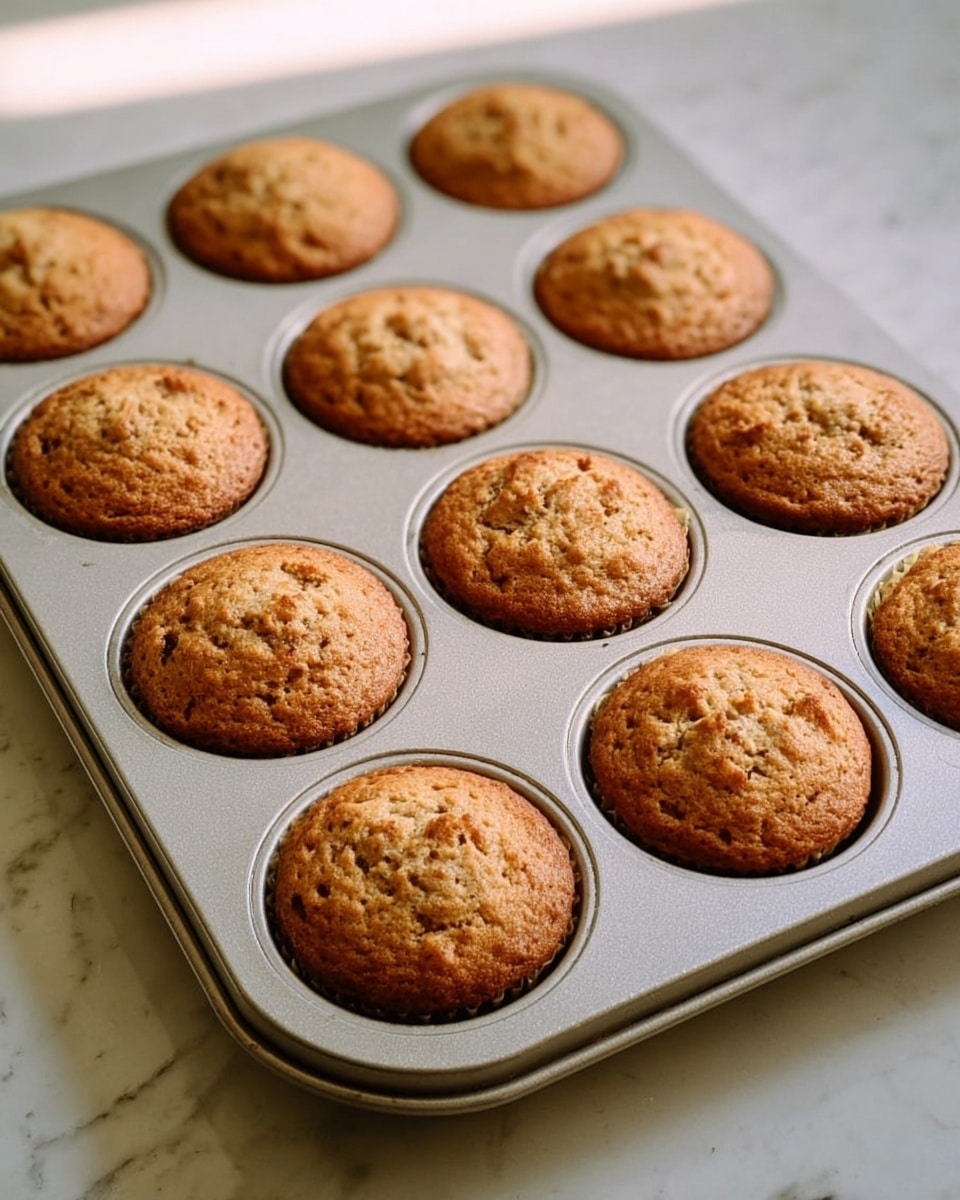 A silver muffin tray filled with twelve evenly baked muffins shown from the top, each muffin is golden brown with slightly cracked tops showing a soft texture inside. The tray rests on a white marbled surface, with the light casting soft shadows around the tray edges. The muffins have a rough surface with small darker brown spots and look warm and freshly baked. photo taken with an iphone --ar 4:5 --v 7