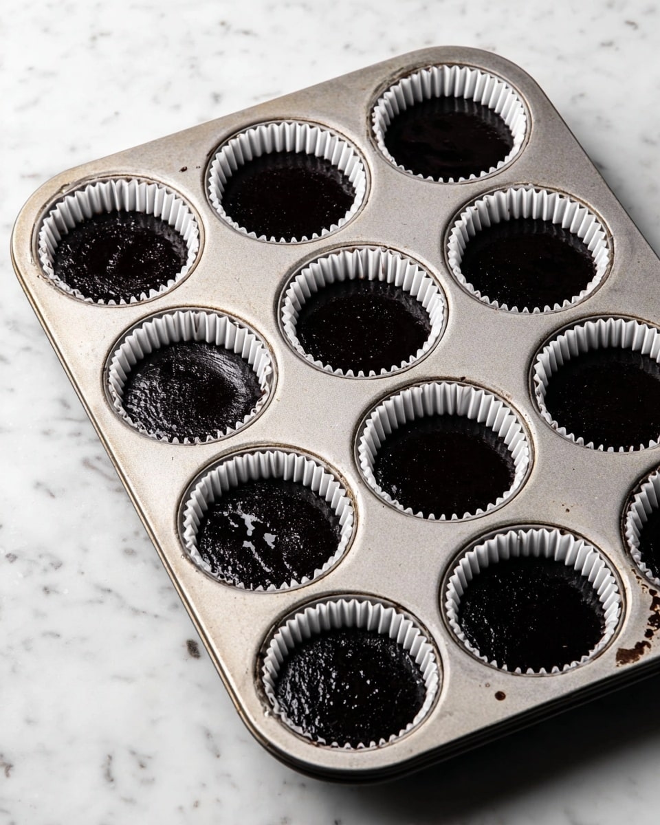A metal muffin tray holds nine white paper liners filled with smooth, black batter that has small bubbles on the surface, indicating it is freshly poured and unbaked. The tray sits on a white marbled surface, with a few small smudges and marks around the edges. The batter fills each liner almost to the top, creating a flat, shiny, and even layer of dark liquid inside each cup. The overall look is clean and simple, showing the preparation stage before baking. photo taken with an iphone --ar 4:5 --v 7