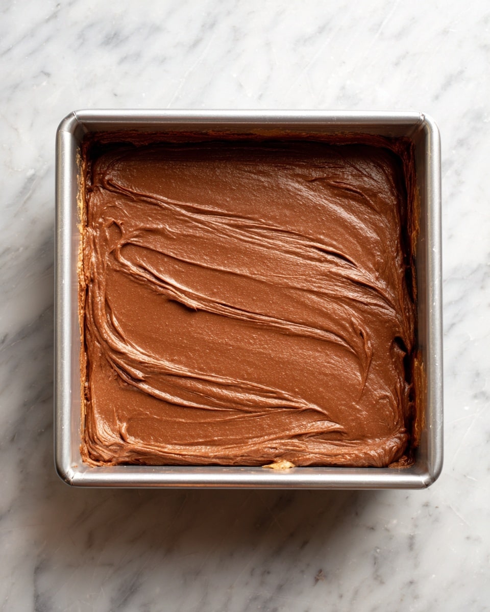 A square metal baking pan shows two layers of chocolate batter. The bottom layer is a lighter brown and smooth, spread evenly across the pan. The top layer is a darker brown, thicker, and spread smoothly over the lighter layer. The pan is placed on a white marbled surface with part of the batter touching the pan edges. There is a small black and white circle with the number 5 on the upper left corner of the image. photo taken with an iphone --ar 4:5 --v 7