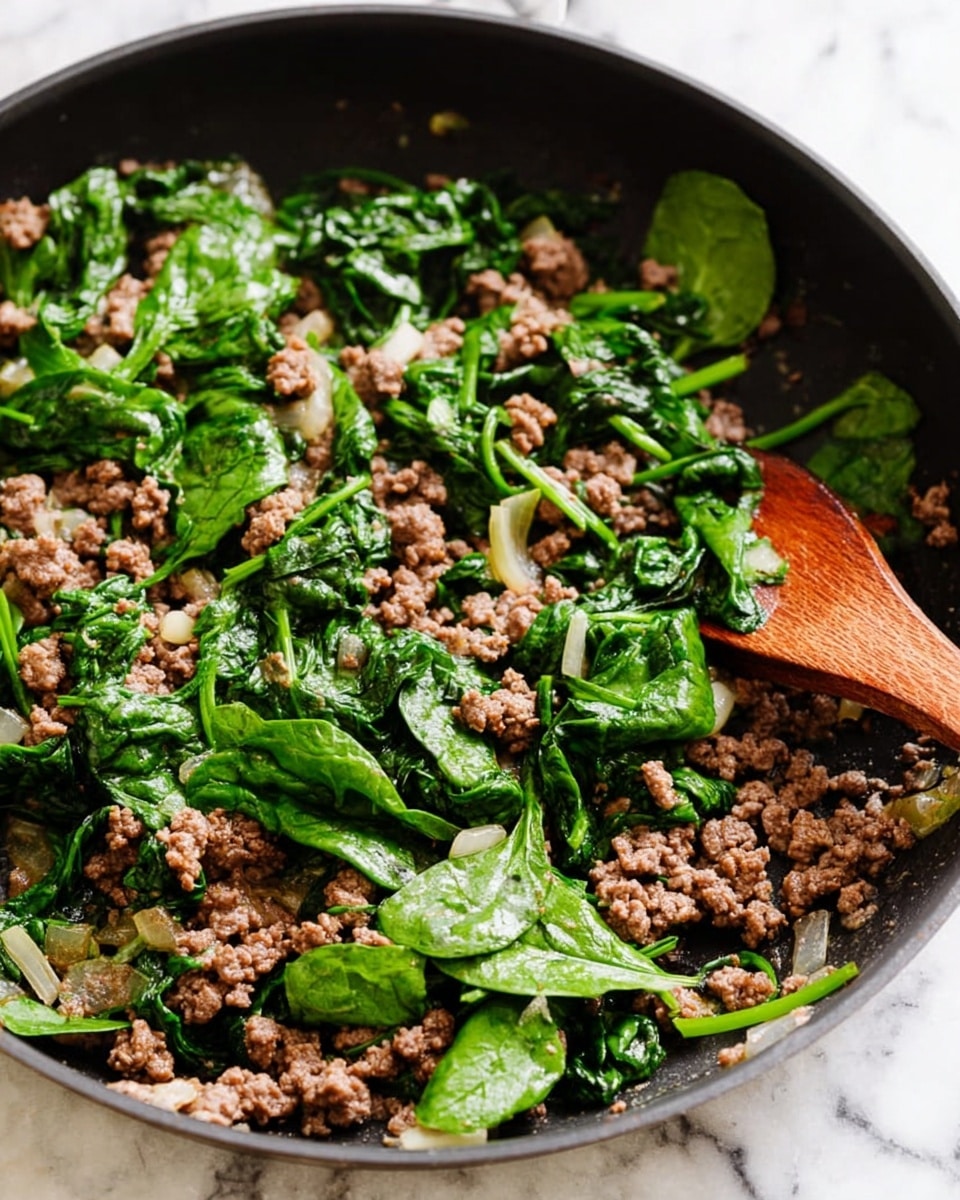 The image shows a close-up of a black frying pan filled with cooked ground beef mixed with bright green spinach leaves and small pieces of chopped onion. The beef is crumbly and brown, while the spinach is fresh and slightly wilted in parts. A wooden spoon rests inside the pan on the right side, partly covered with some spinach leaves and beef bits. The background is a white marbled surface. photo taken with an iphone --ar 4:5 --v 7