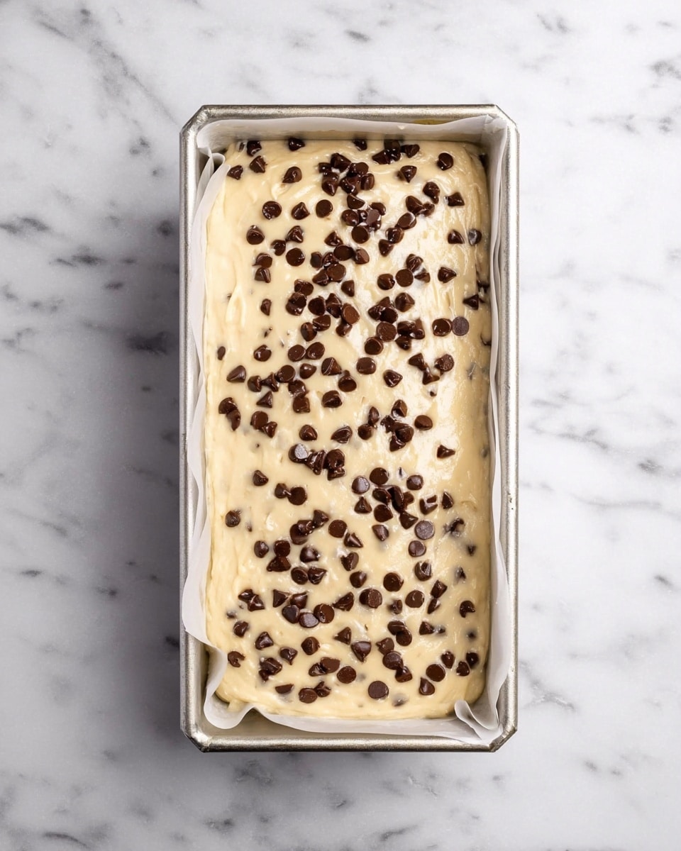 A metal rectangular loaf pan lined with white parchment paper holds a thick, creamy beige batter filled with small dark chocolate chips spread throughout. The top of the batter is smooth but slightly uneven, generously sprinkled with more dark chocolate chips scattered in a random pattern. The pan is placed on a white marbled surface that adds a clean and bright feel to the image. photo taken with an iphone --ar 4:5 --v 7