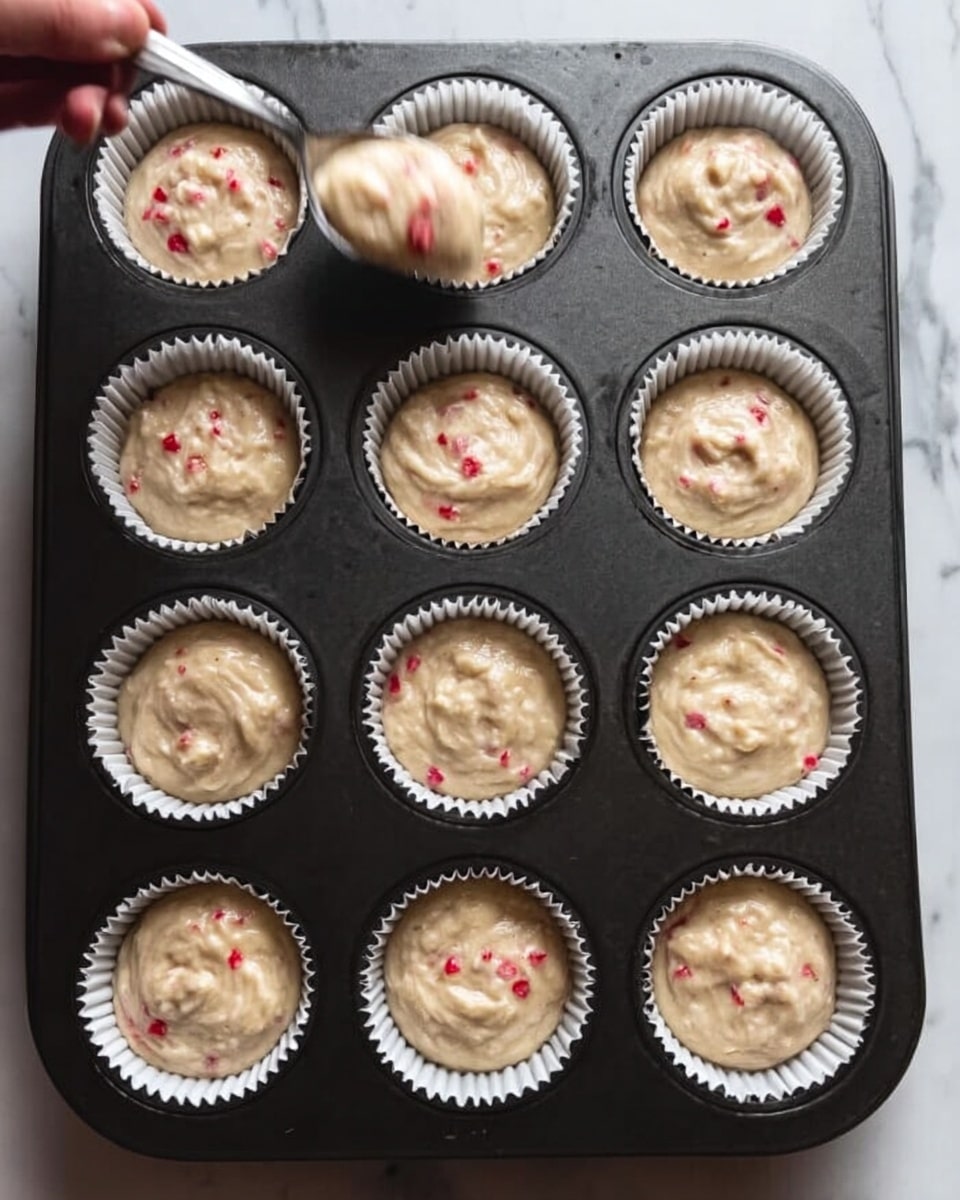 A dark baking tray with twelve white cupcake liners filled with light beige batter that has small red bits mixed in. The batter is loosely swirled, filling the liners just over halfway. A woman's hand holds a spoon above the tray, appearing to drop more batter into one of the liners. The surface below the tray is a white marbled texture. photo taken with an iphone --ar 4:5 --v 7