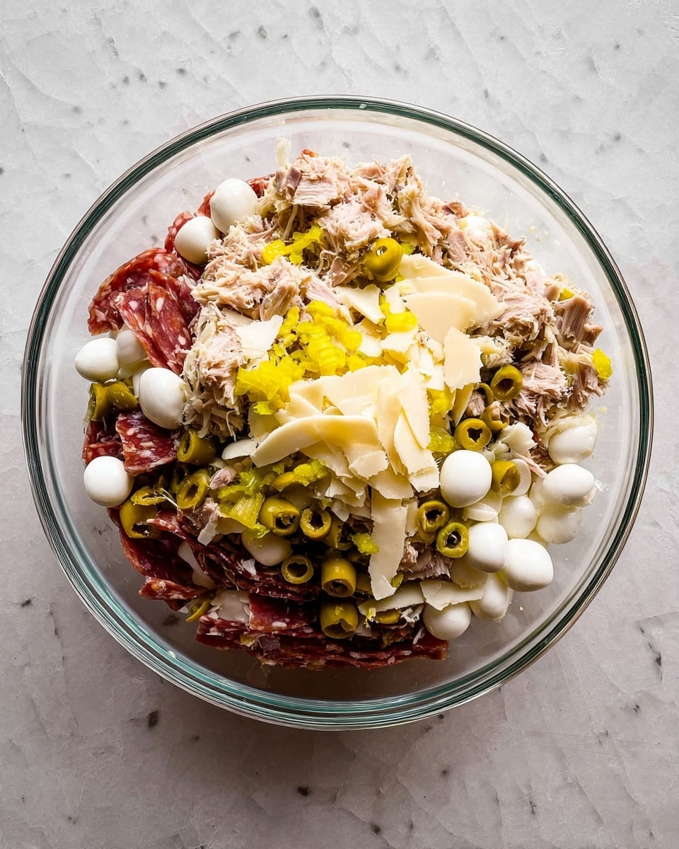 The image shows a clear glass bowl filled with several layers of ingredients on a white marbled surface. The bottom layer consists of thinly sliced reddish-brown cured meat. On top of that, there are small white mozzarella balls, scattered green olive slices, and yellow pickled pepper rings mostly placed around the edges. Above these layers, there are shredded light brown tuna pieces mixed with small flat pale yellow cheese slices scattered on top. The mix creates a colorful, textured contrast with the white marbled background. photo taken with an iphone --ar 4:5 --v 7