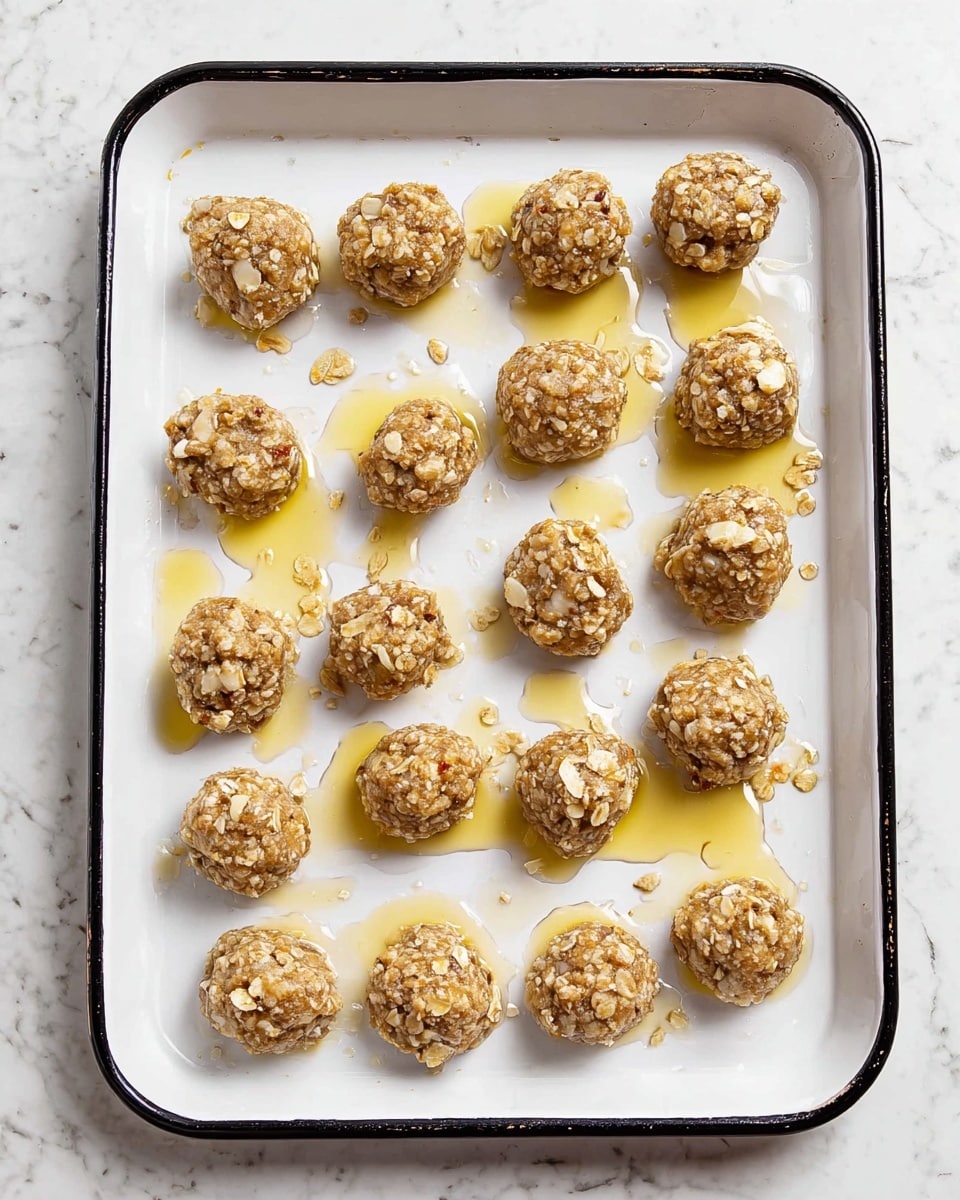 The image shows a white baking tray with around 30 small, round cookie dough balls placed evenly in rows. Each ball is light brown with visible white chunks mixed inside and has a rough texture that looks like it contains oats or nuts. Some spots have a golden shine indicating melted butter or syrup, giving a slightly sticky look around the dough balls. The tray is lined with white parchment paper, and the tray sits on a white marbled surface. The scene is well-lit with a clean and simple background. photo taken with an iphone --ar 4:5 --v 7
