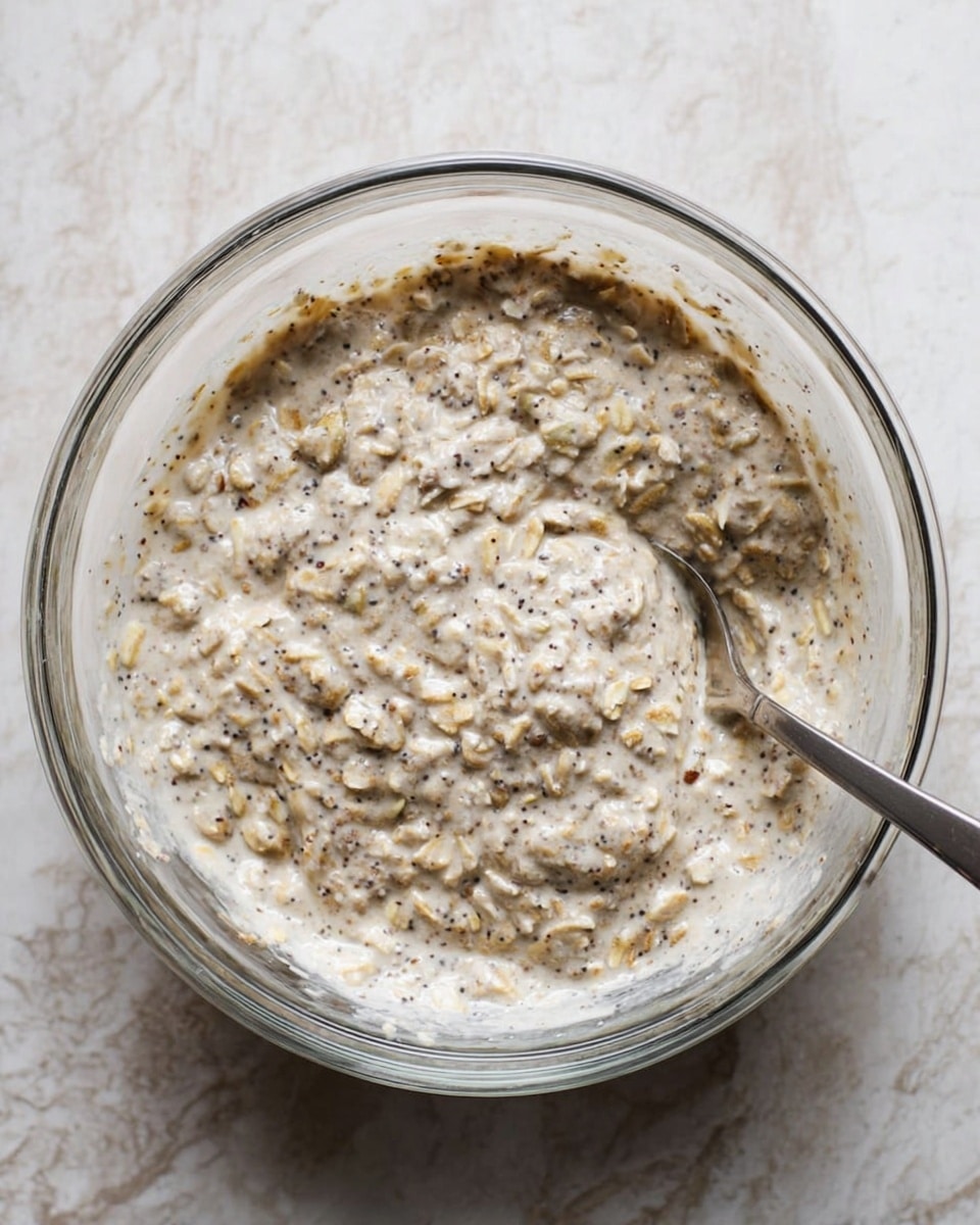 There is a clear glass bowl placed on a white marbled surface, filled with a creamy mixture of oats and seeds. The mixture is light beige with small dark specks evenly spread throughout, giving it a textured look. The oats are soft and chunky, blending smoothly with the creamy base, while the small specks add contrast. A metal spoon is partially dipped in the bowl, resting on the right side inside the mixture. The overall appearance is thick and hearty, with a rustic, natural feel. Photo taken with an iphone --ar 4:5 --v 7