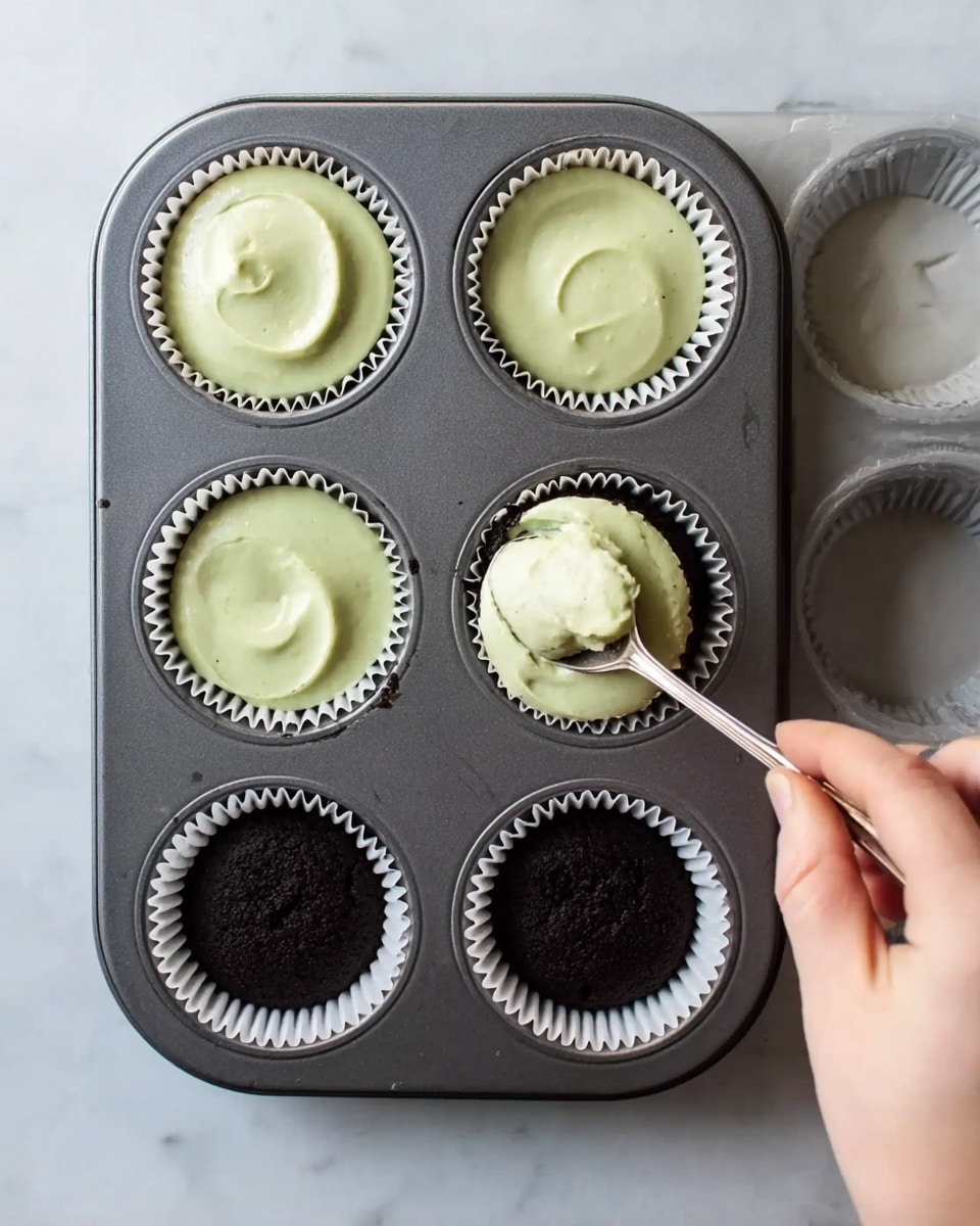 A dark gray metal muffin tray holds six white paper liners, some filled with a smooth, pale green batter. The bottom three liners have a dark crumb base, two of them topped with the green batter. On the right, a woman's hand holds a silver spoon scooping the pale green batter into one of the liners with the dark crumb base. The tray rests on a white marbled surface, and the lighting is soft and natural. photo taken with an iphone --ar 4:5 --v 7