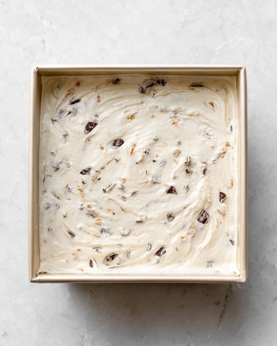 A square baking pan filled with a smooth, creamy white mixture that has small, unevenly scattered dark chocolate chunks and light brown bits mixed in. The surface of the mixture is softly textured with gentle swirls and slight peaks, filling the pan evenly and reaching the edges. The pan sits on a white marbled surface with a clean, bright background. Photo taken with an iphone --ar 4:5 --v 7