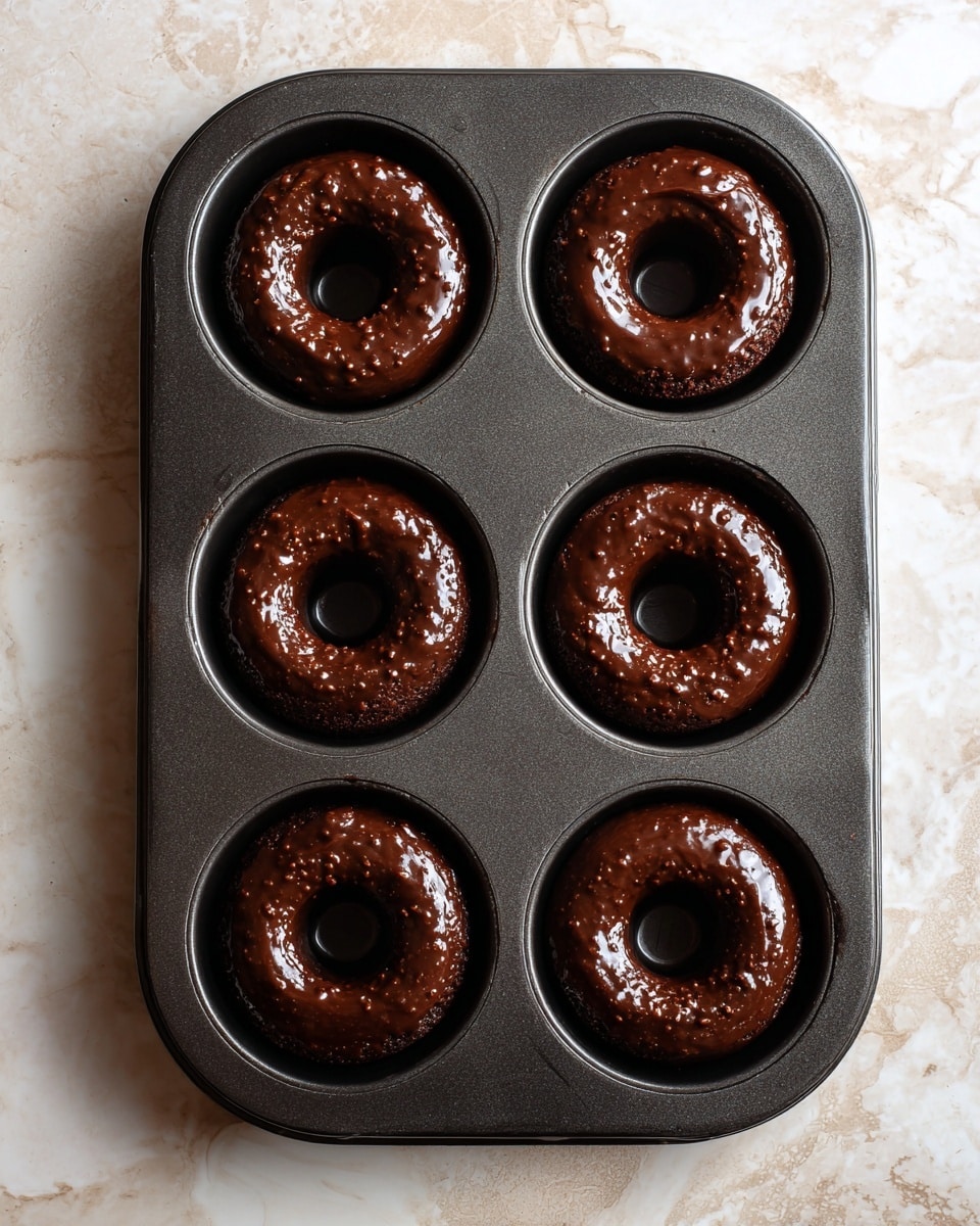 Two side-by-side images show a dark non-stick donut baking tray with six donuts each on a white marbled surface. The left image shows the tray filled with raw chocolate batter, shiny and thick with small lumps, filling each of the six donut molds evenly. The right image shows the same tray after baking, with six smooth, round, dark chocolate donuts, each with a defined hole in the center and a soft cracked surface texture. The tray is centered on the white marbled surface in both images photo taken with an iphone --ar 4:5 --v 7