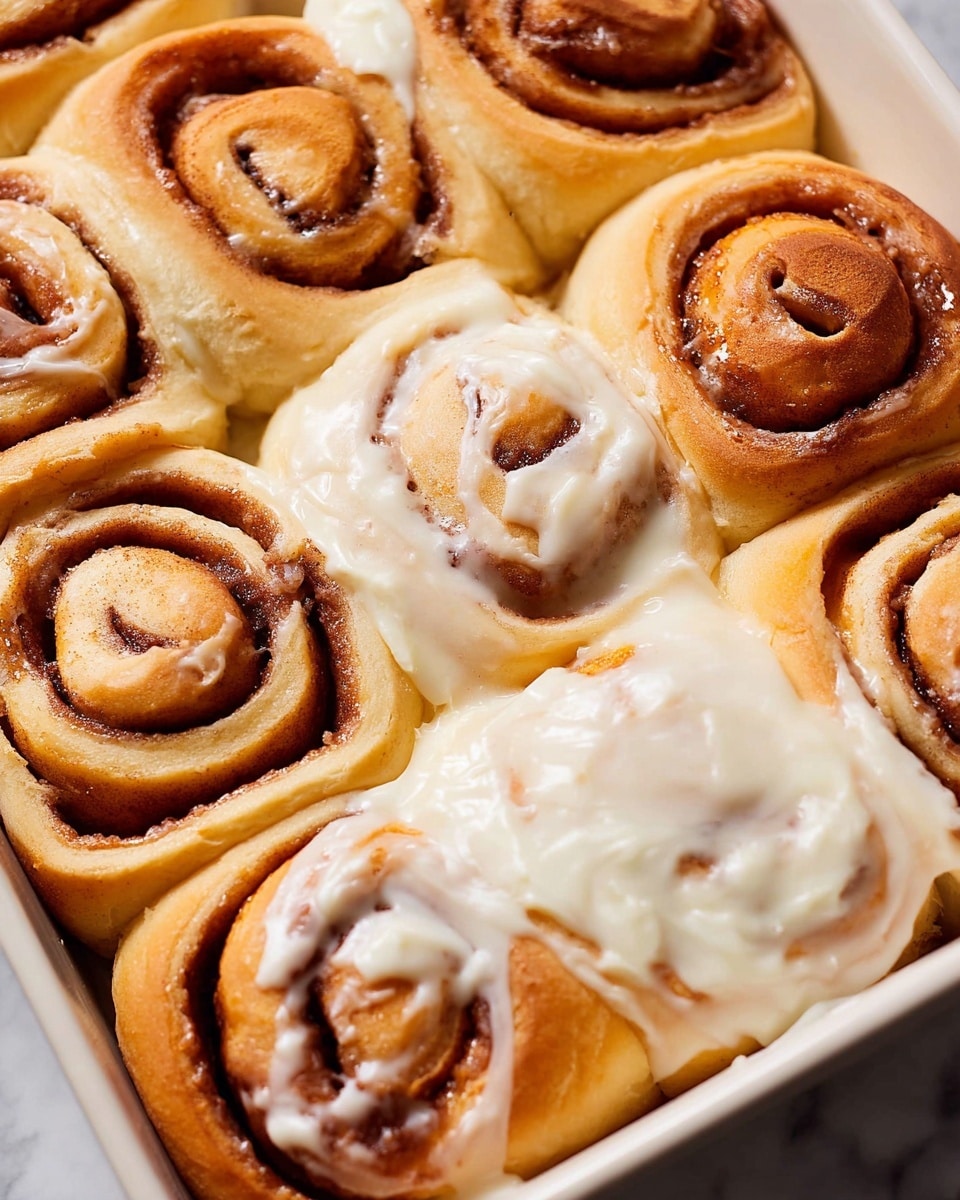 The image shows a close-up of a white baking dish filled with soft, golden brown cinnamon rolls. The rolls are arranged tightly together, with visible layers of dough spiraled around a rich cinnamon filling that is darker brown and slightly shiny. Some rolls are covered with a thick layer of creamy, white icing that has a smooth texture, partially melting and dripping onto the rolls beneath. The overall look is warm and inviting, with the contrast between the warm golden dough and the cool white icing standing out against the white marbled surface. photo taken with an iphone --ar 4:5 --v 7
